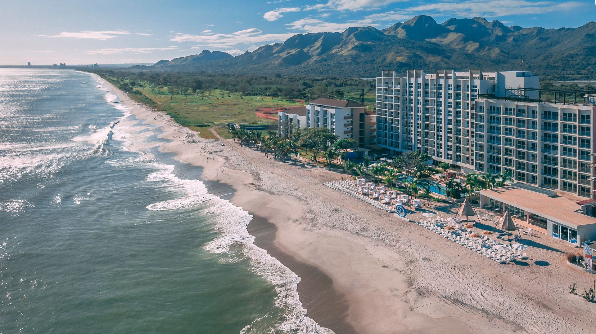 Ocean view of a beach with waves, sand, buildings, and mountains in the background under a blue sky.
