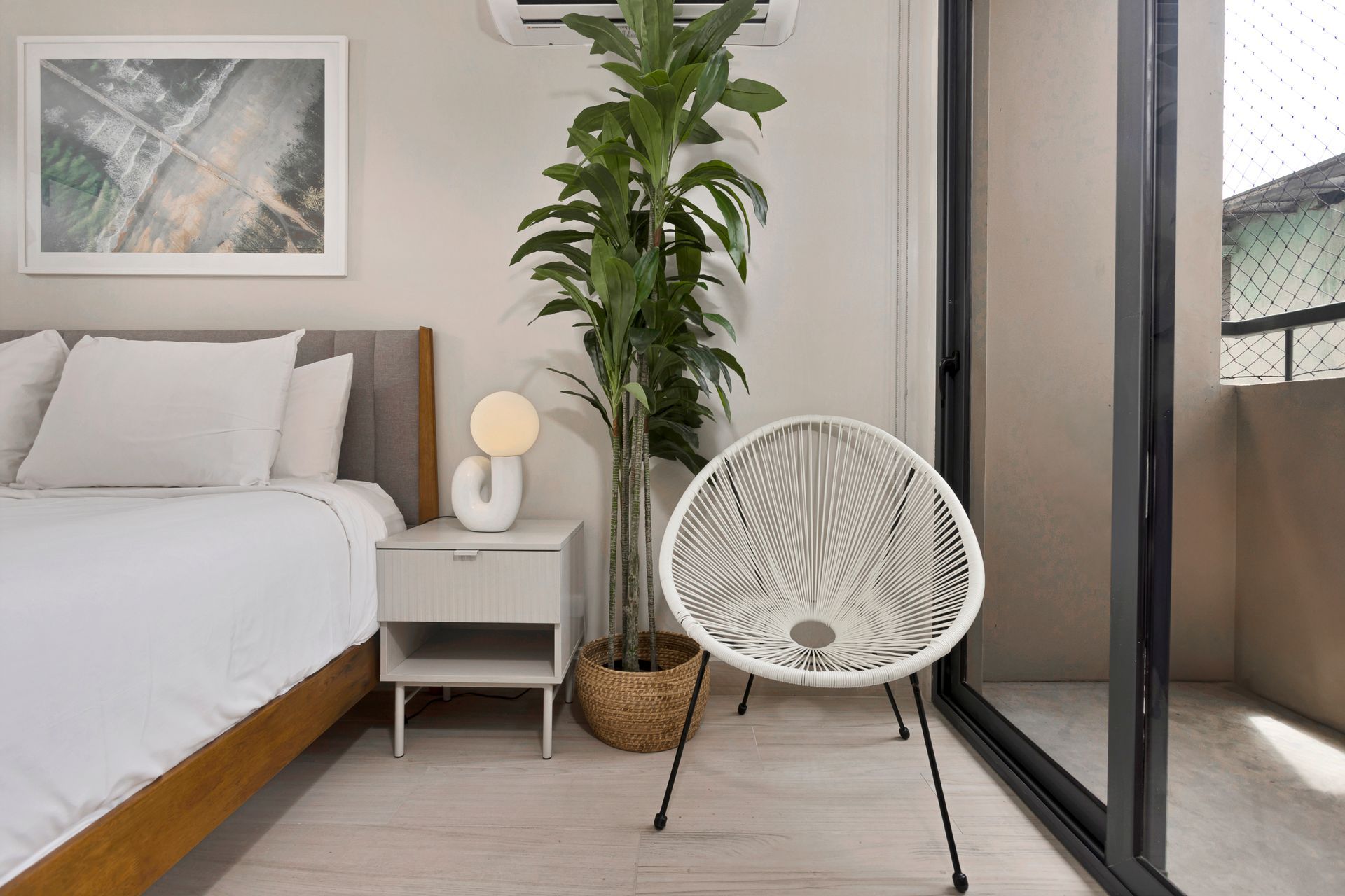 Bedroom with bed, nightstand, tall potted plant, white woven chair, and balcony.
