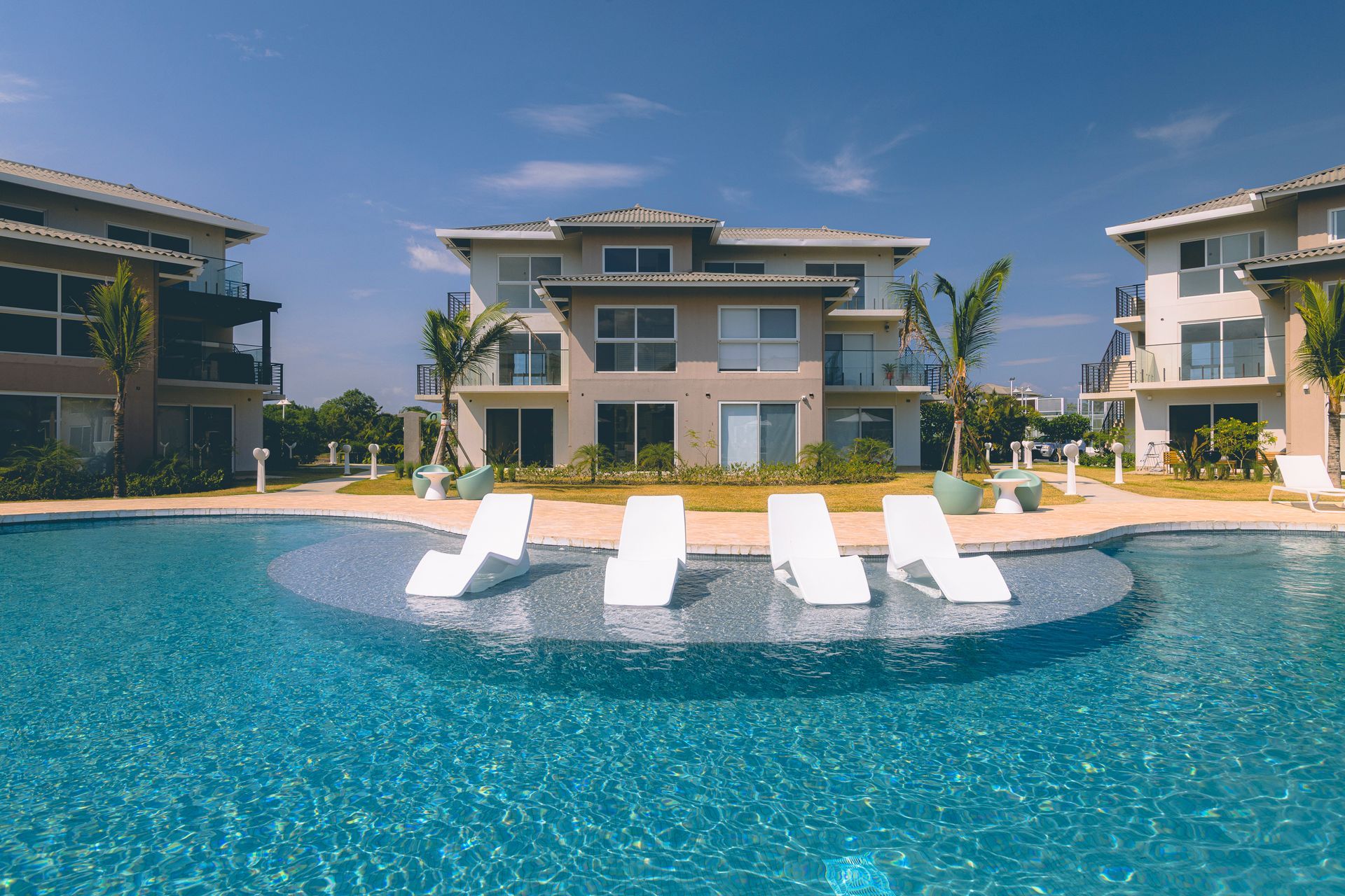 Pool with white lounge chairs in front of multi-story buildings on a sunny day.