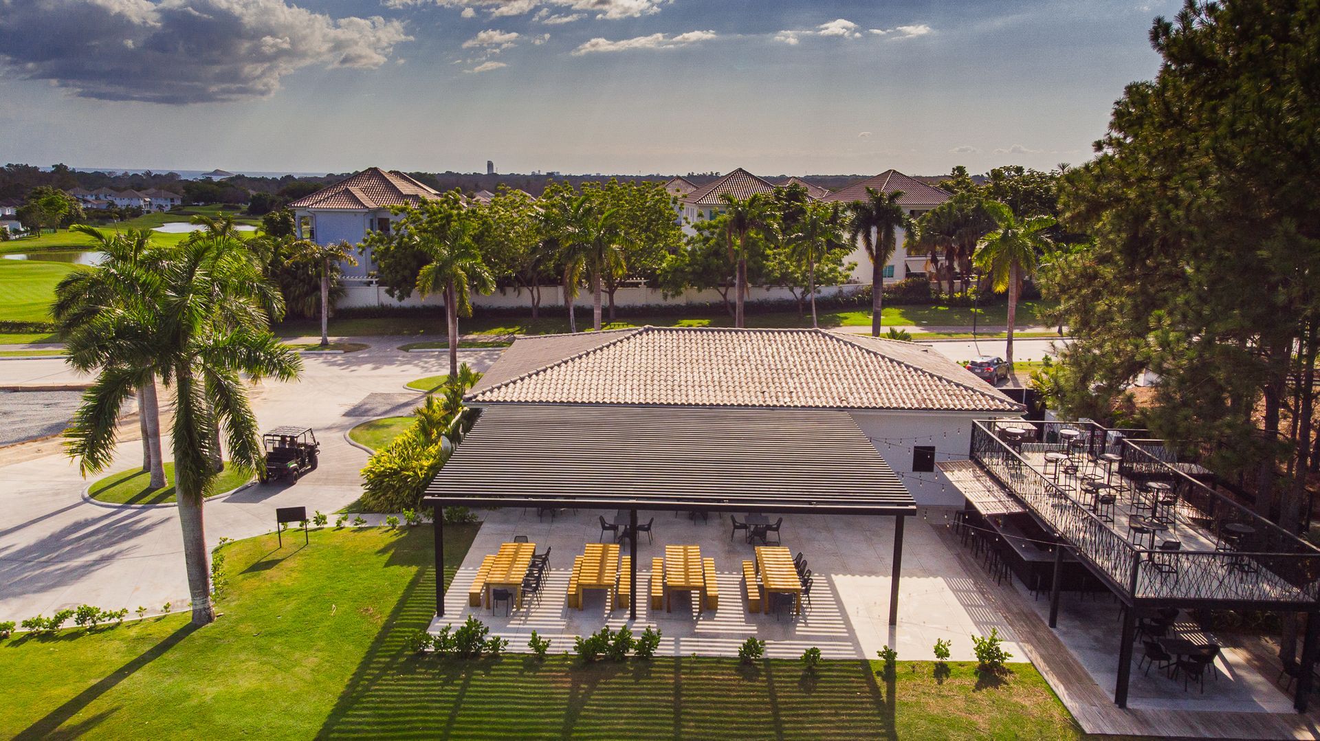 Aerial view of a patio with lounge chairs. A building with a wavy awning. Palm trees and green grass are nearby.