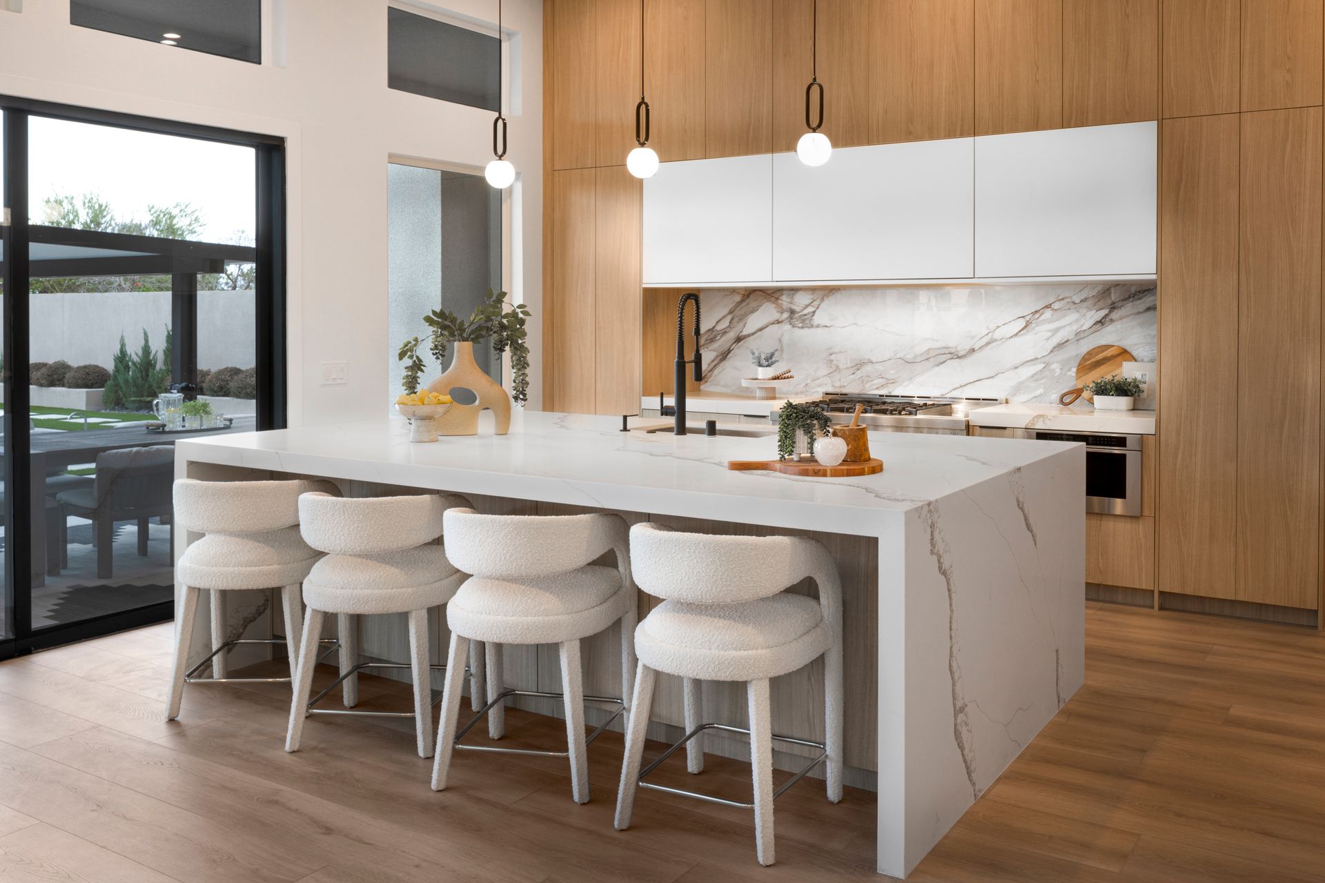 Modern kitchen with white island, wood cabinets, and marble backsplash. Five white bar stools.