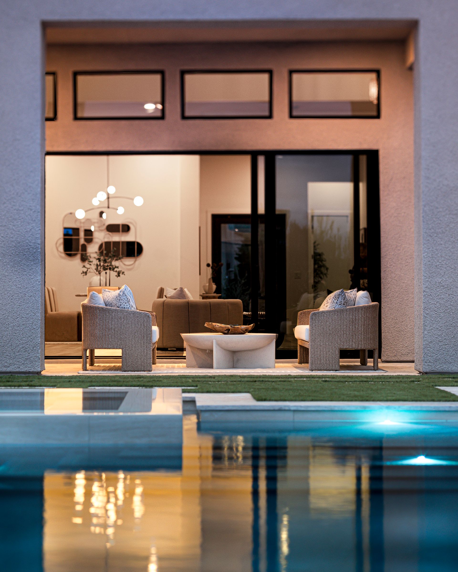 Outdoor view into a modern home, showcasing a living room with pool reflection and patio seating.