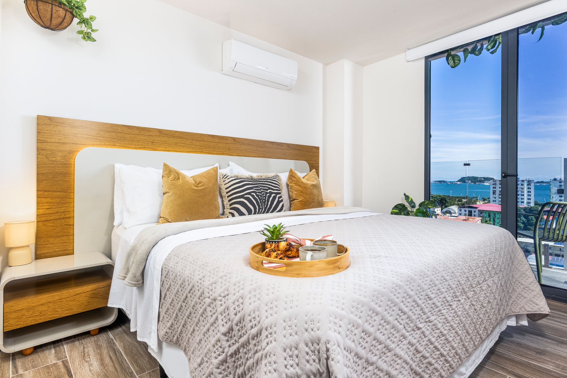 Bedroom with king-size bed, tray of food, large window with balcony, white walls, and wooden accents.