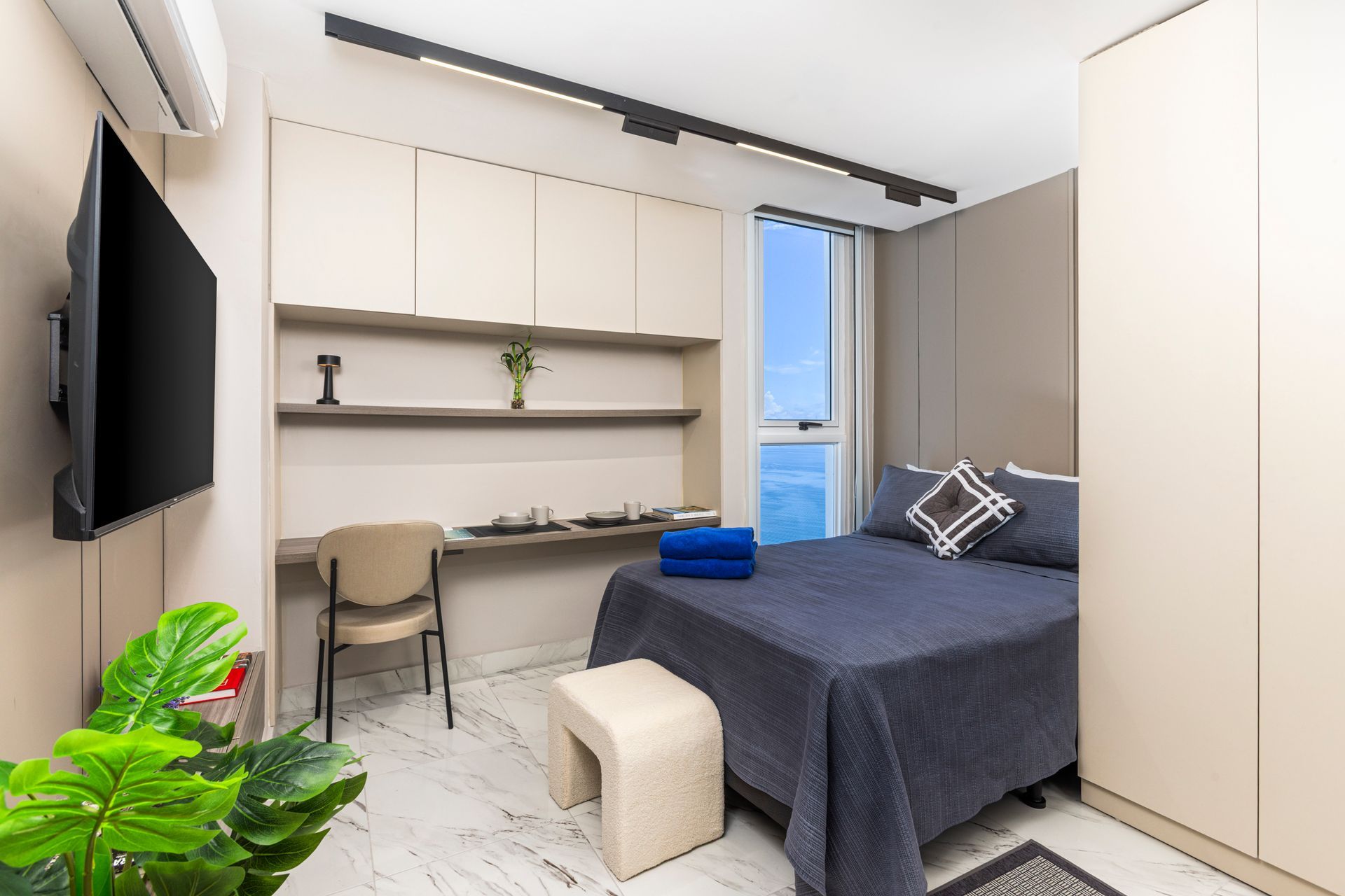 A modern, compact bedroom with built-in desk, bed, and small window with ocean view.