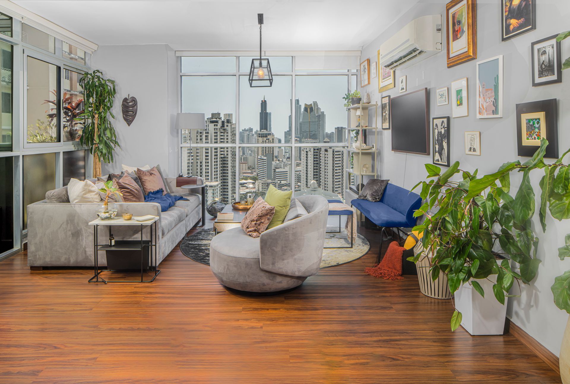 Living room with city view, grey sofa, accent chairs, artwork, plants, and hardwood floors.