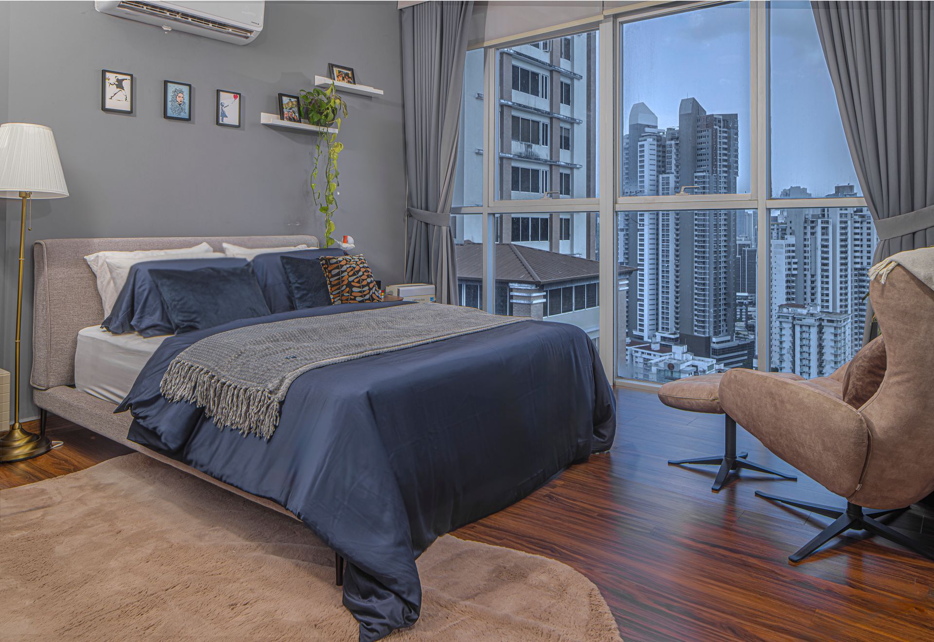 Bedroom with large window overlooking city; gray walls, blue bedding, brown chair.