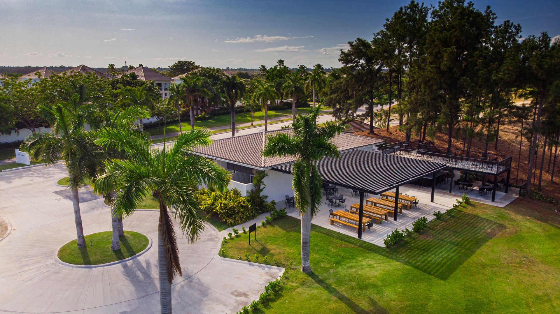 Aerial view: outdoor dining area with tables under a striped pergola, palm trees, and green grass.
