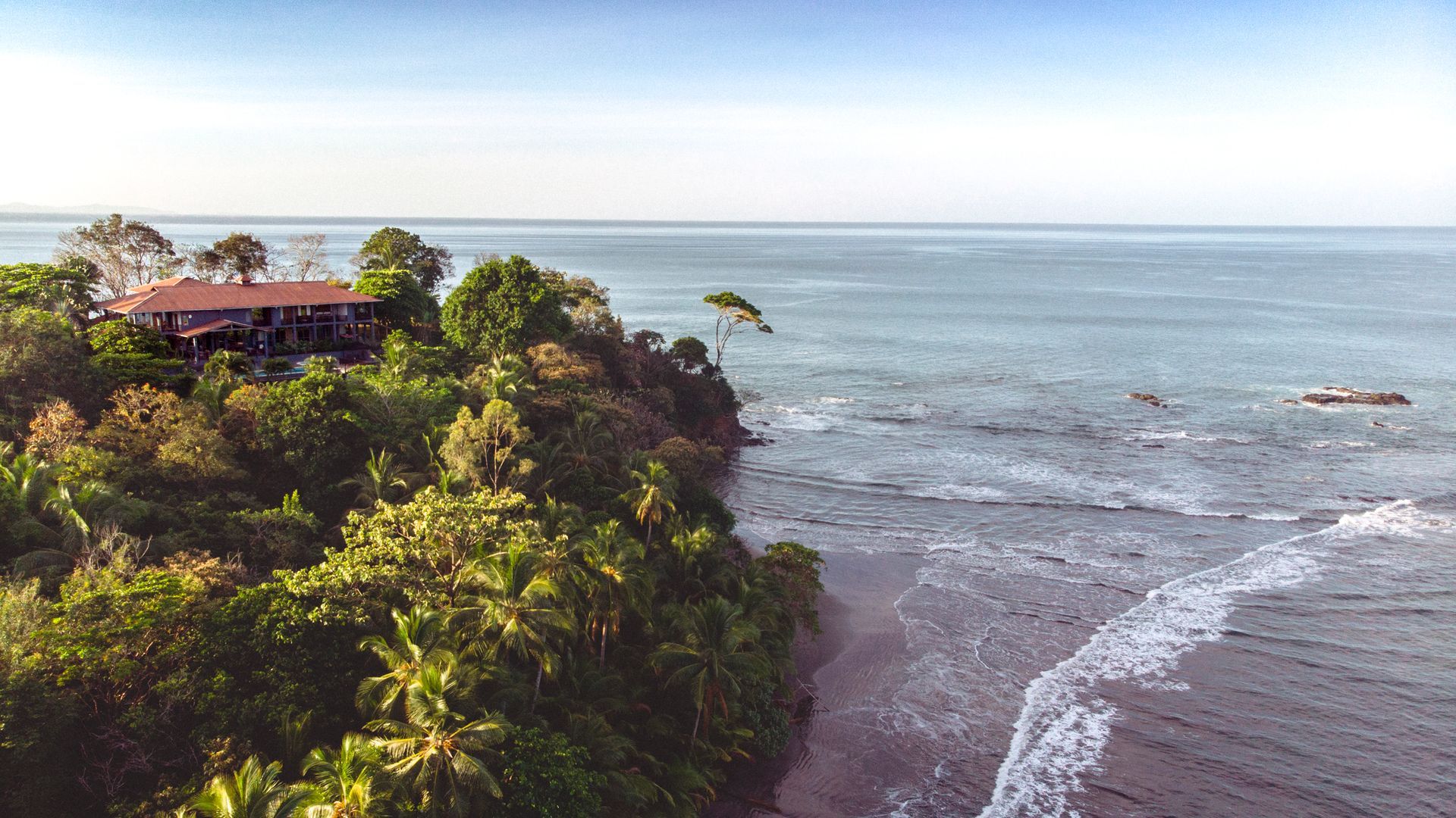 House on a cliff overlooking the ocean with lush green vegetation and a clear blue sky.