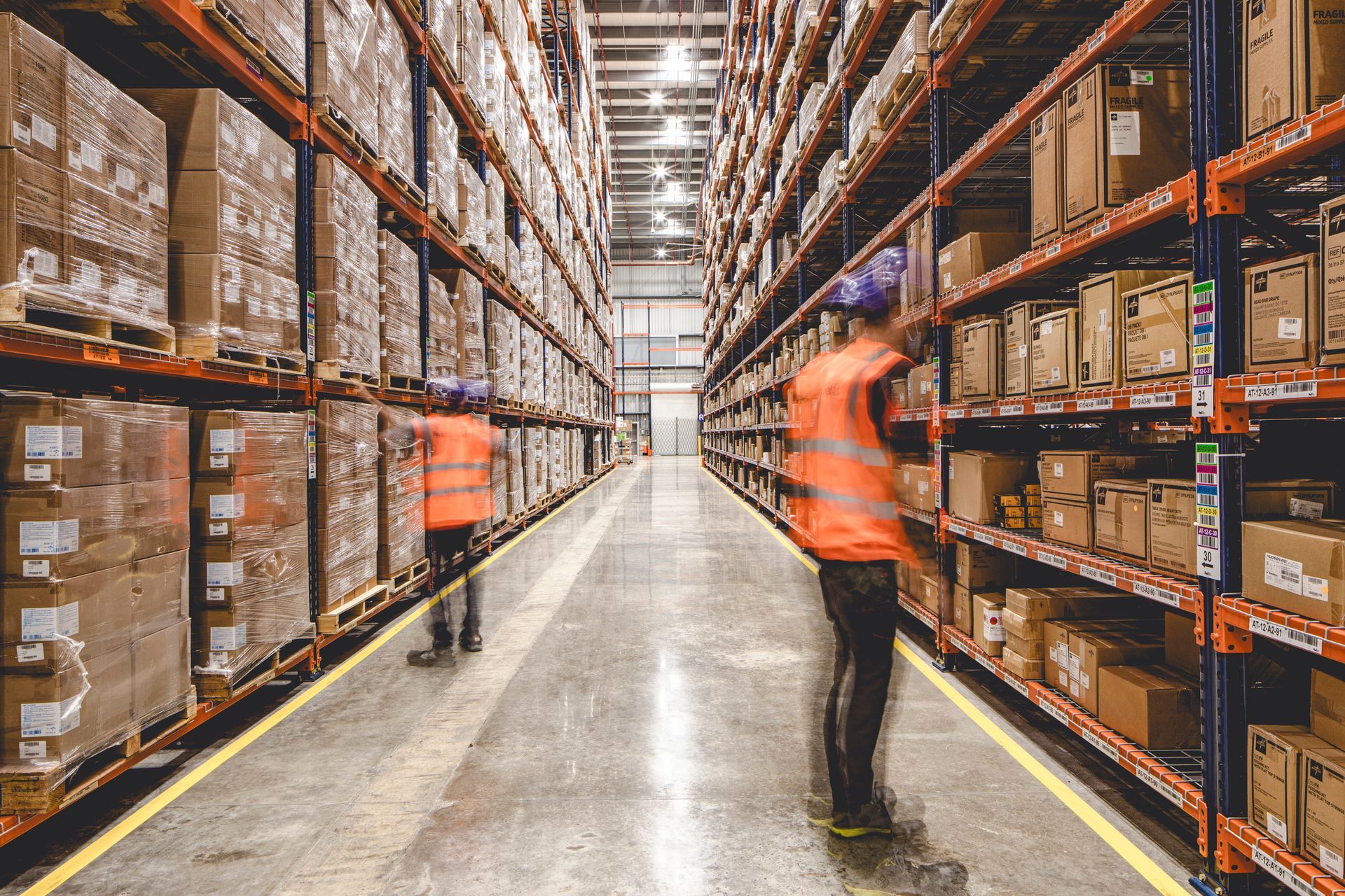 Warehouse interior with workers, high shelves full of boxes, fluorescent lights.