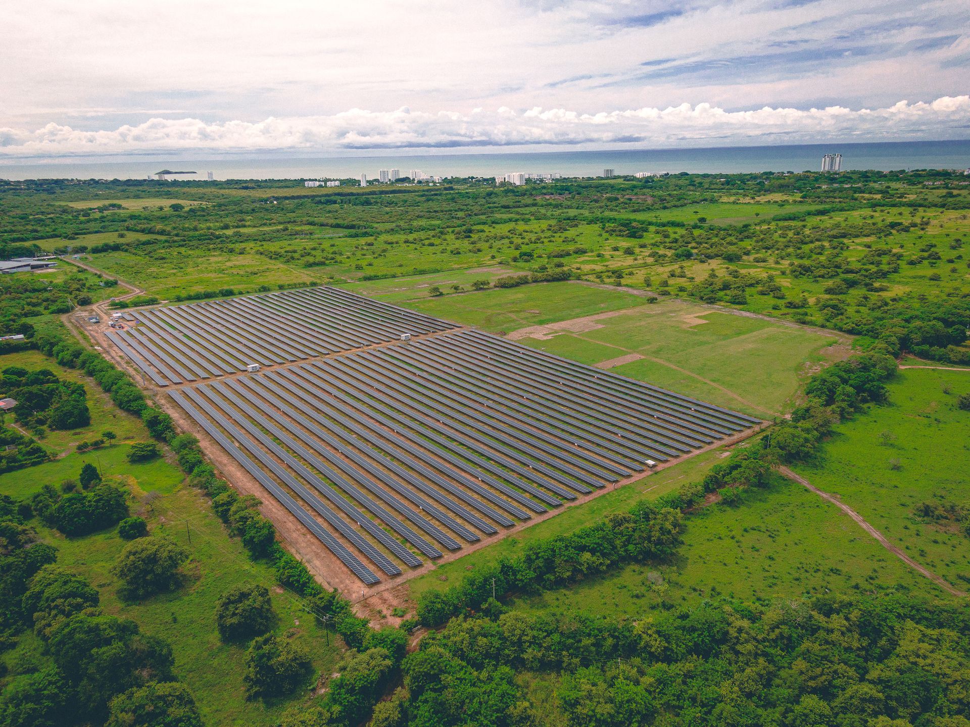 Aerial view of a solar farm in a field with coastline and cloudy sky in the background.
