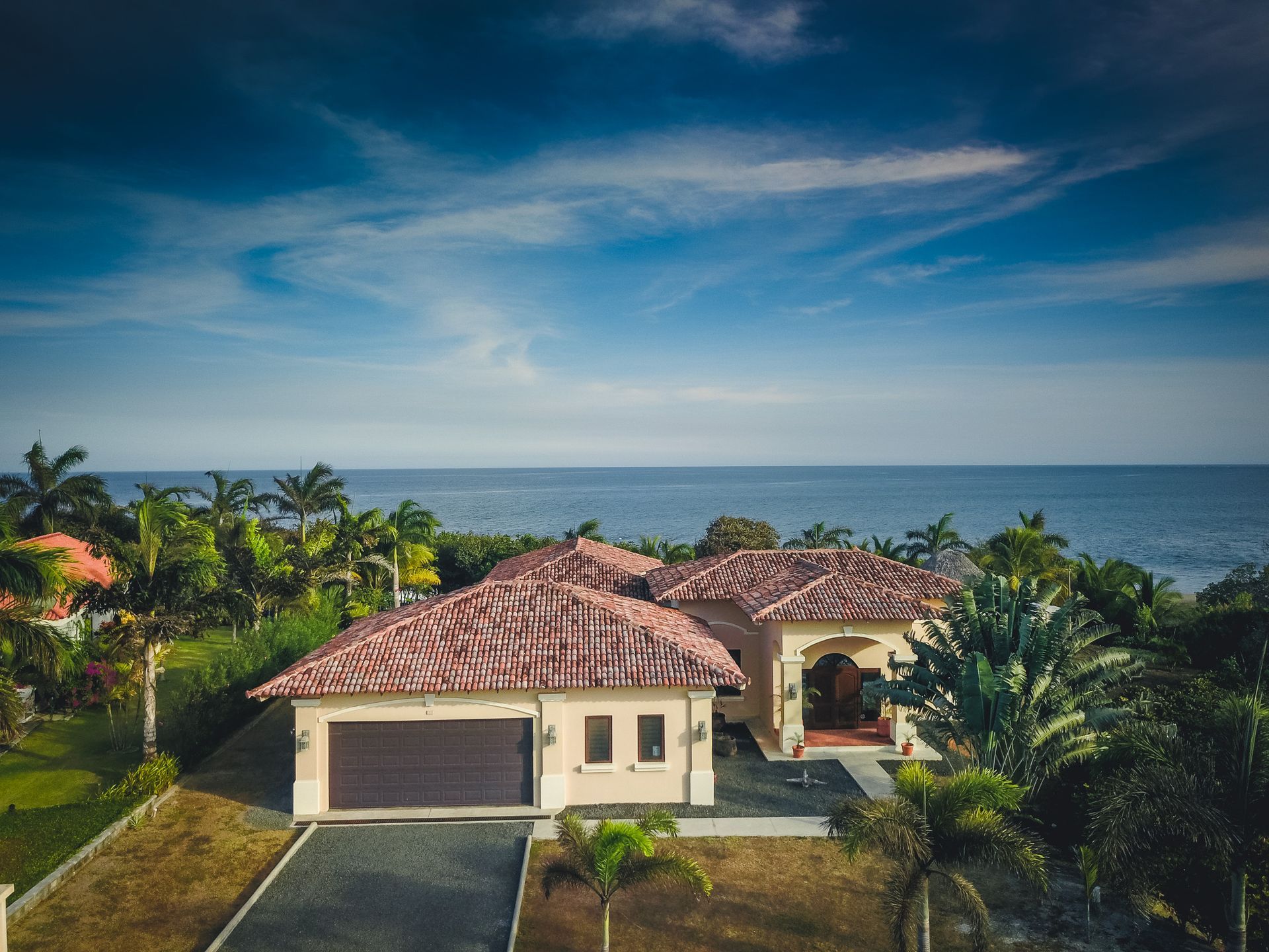 Coastal home with red tile roof, surrounded by green trees, with ocean view under a blue sky.