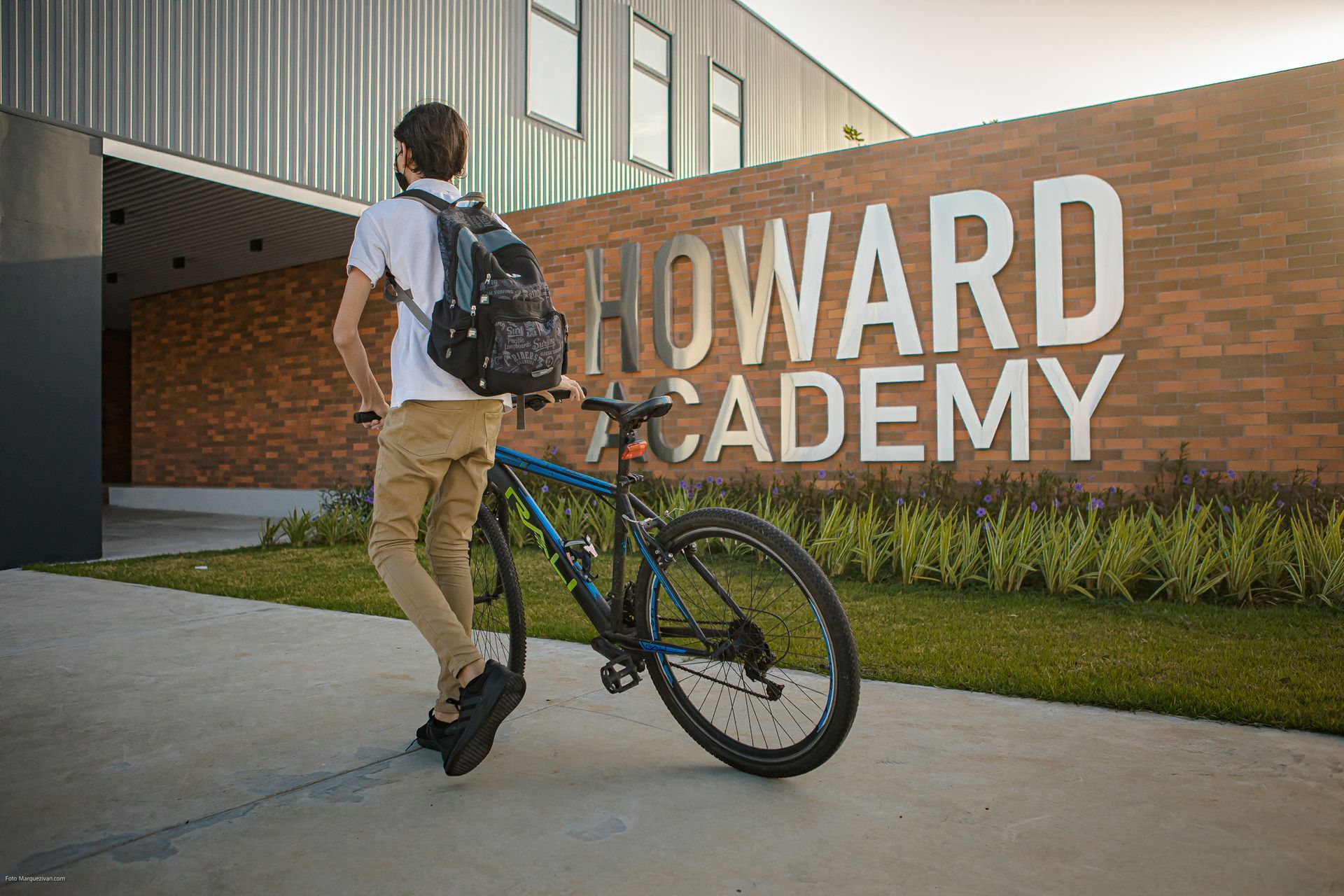 A student with a backpack and bike stands outside Howard Academy, a brick building with a sign.