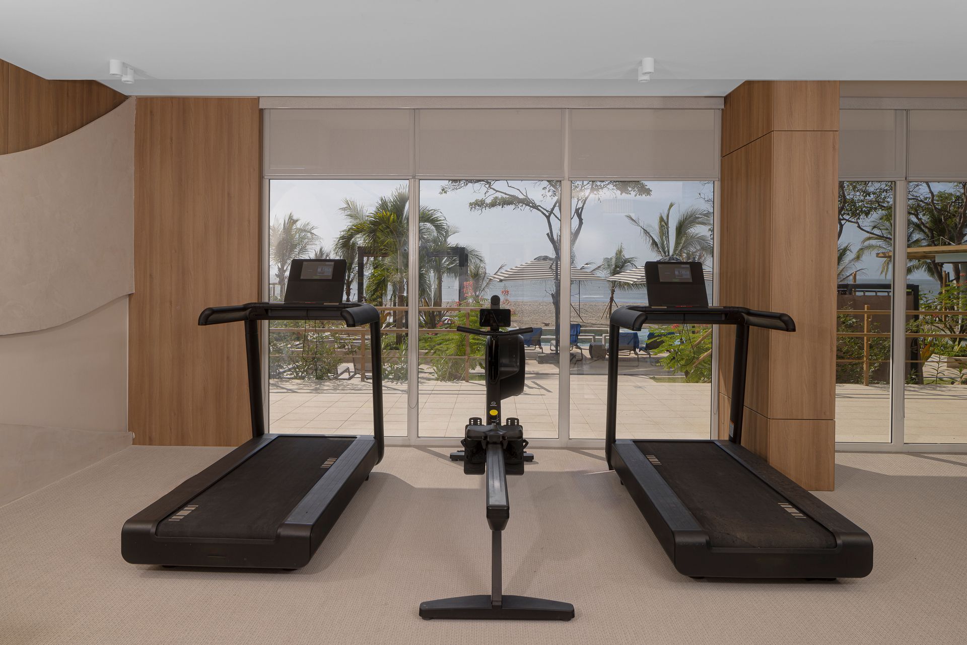 Gym with treadmills and rowing machine, facing a view of the beach and palm trees.
