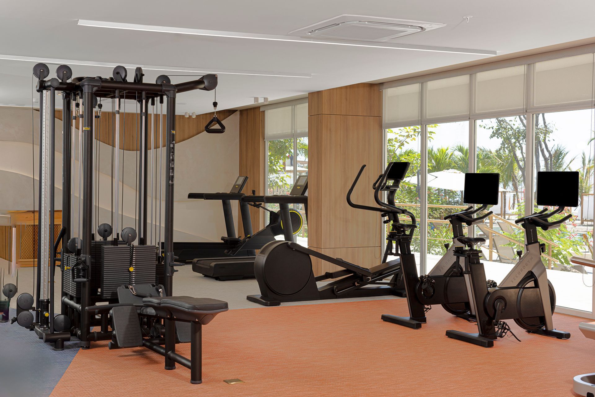 Gym with weight machine, treadmills, elliptical, stationary bikes, and large windows. Orange and wood accents.