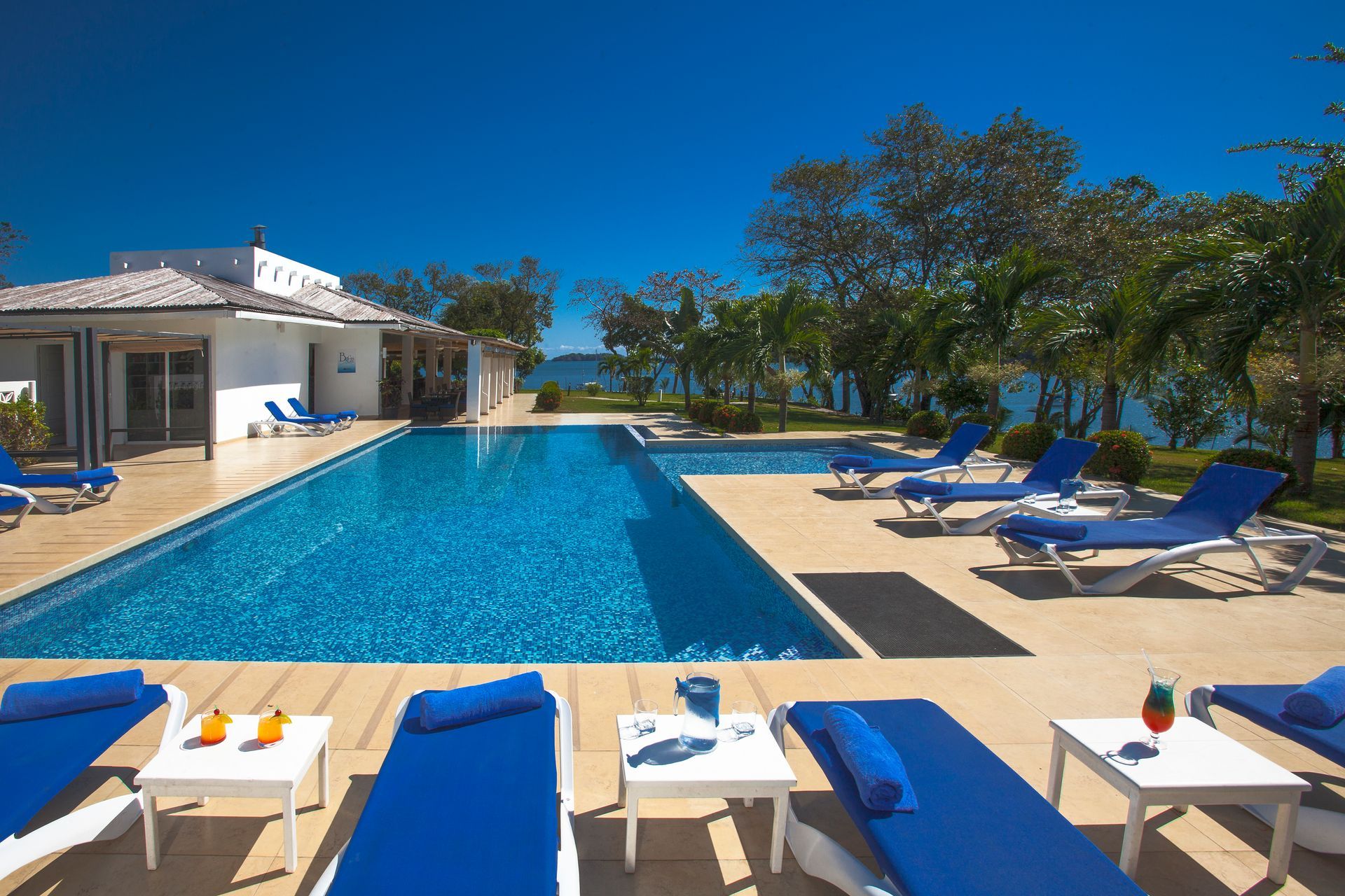 Poolside scene: blue pool, lounge chairs, white tables, clear sky, near water.