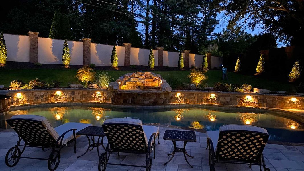 A swimming pool is lit up at night with chairs and tables around it.