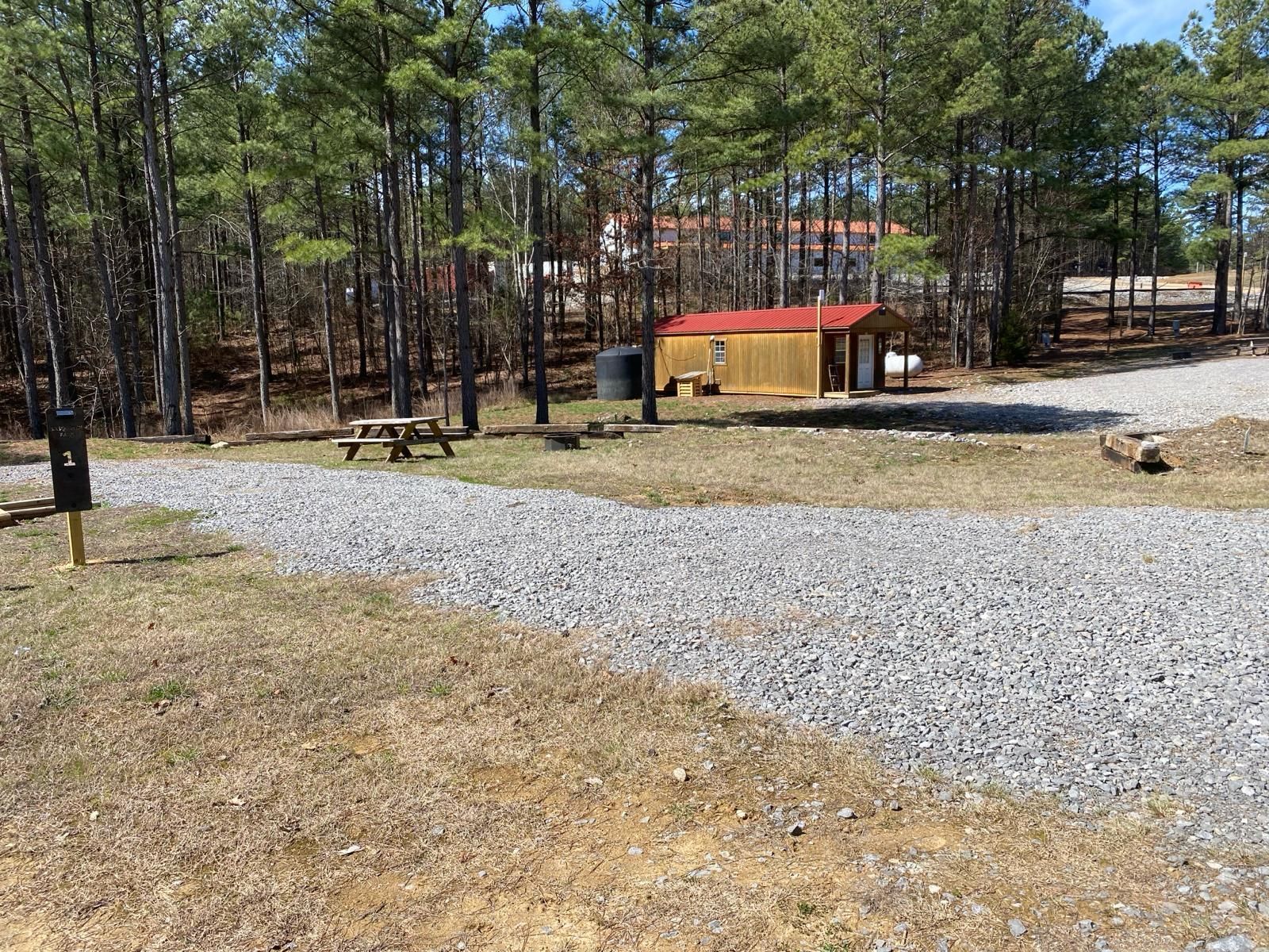 A wooden shed with a red roof is sitting in the middle of a gravel lot in the middle of a forest.