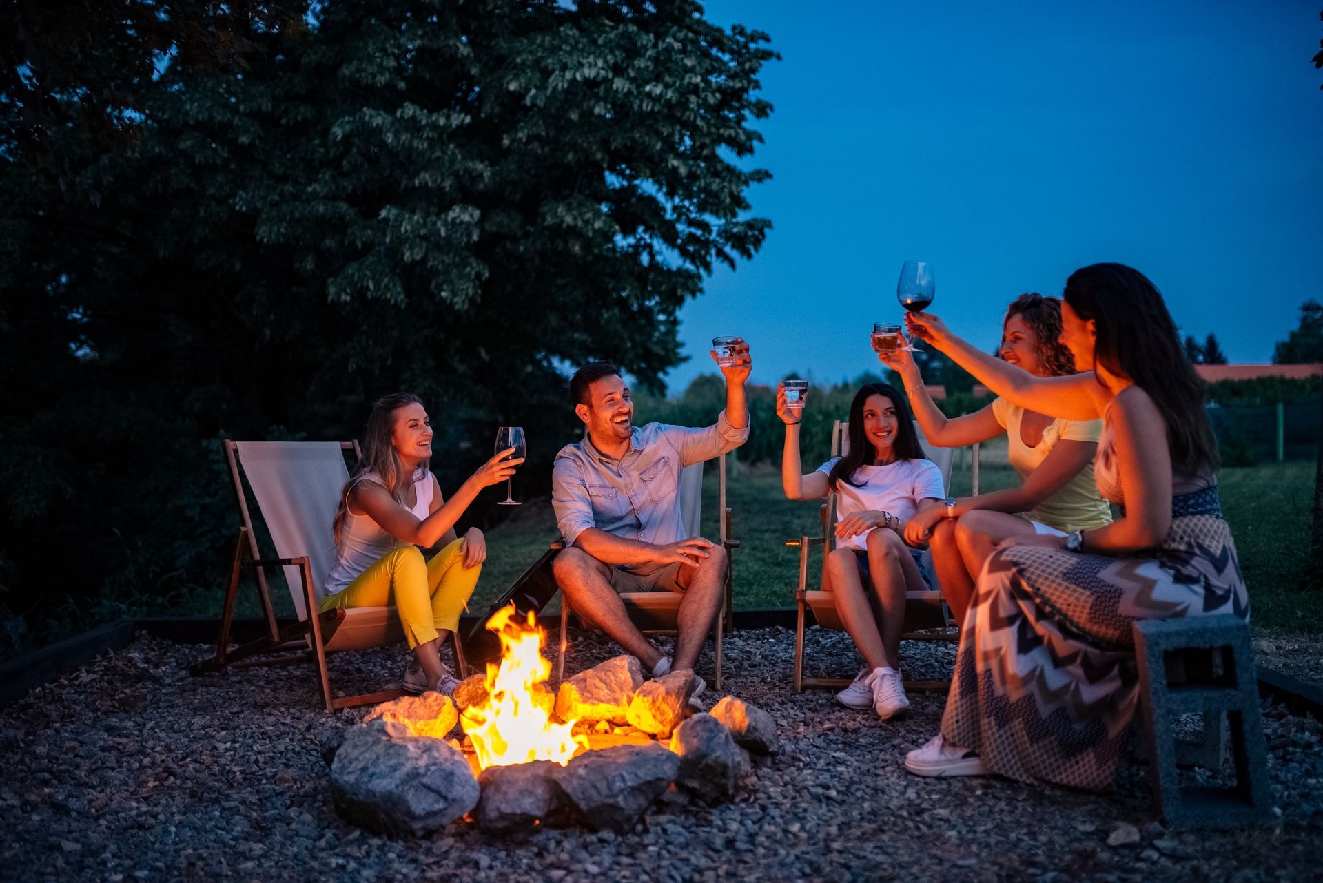 Friends gather around a bonfire at dusk, raising glasses in a toast. People are smiling and laughing in a backyard.
