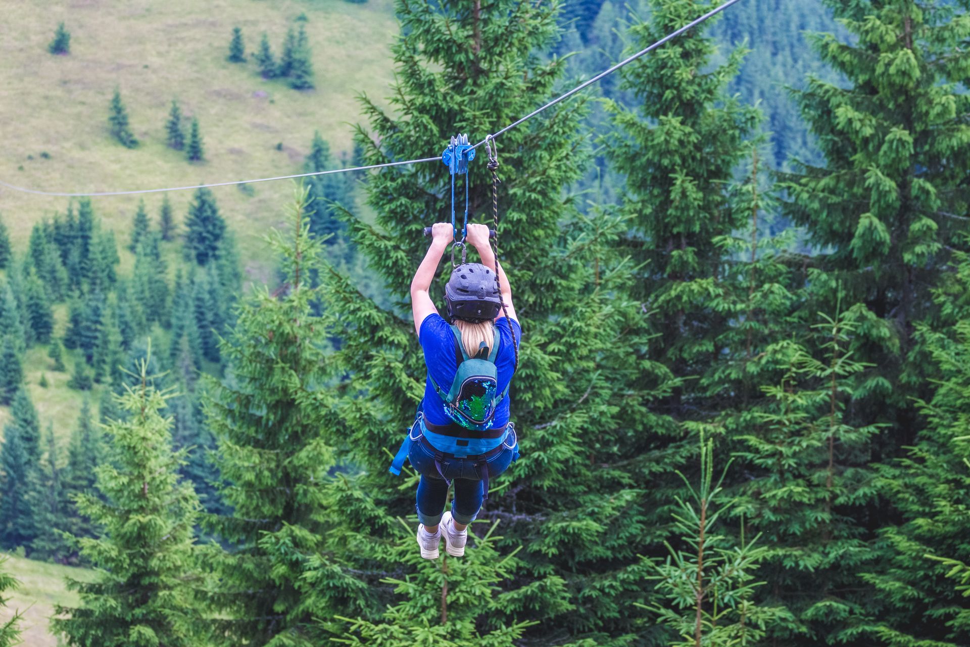 Person ziplining through a forest, seen from the back. They wear a helmet and backpack and are suspended in the air.