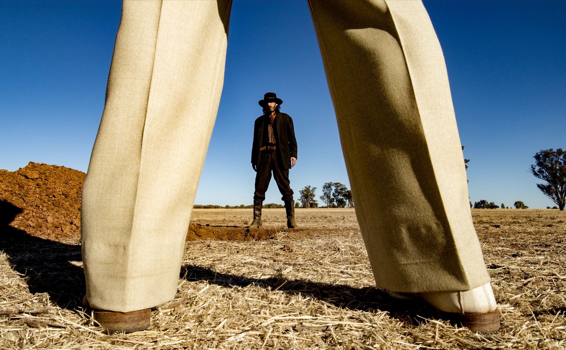Cowboy in a black coat and hat stands in the desert; wide shot from person's legs, beige pants.