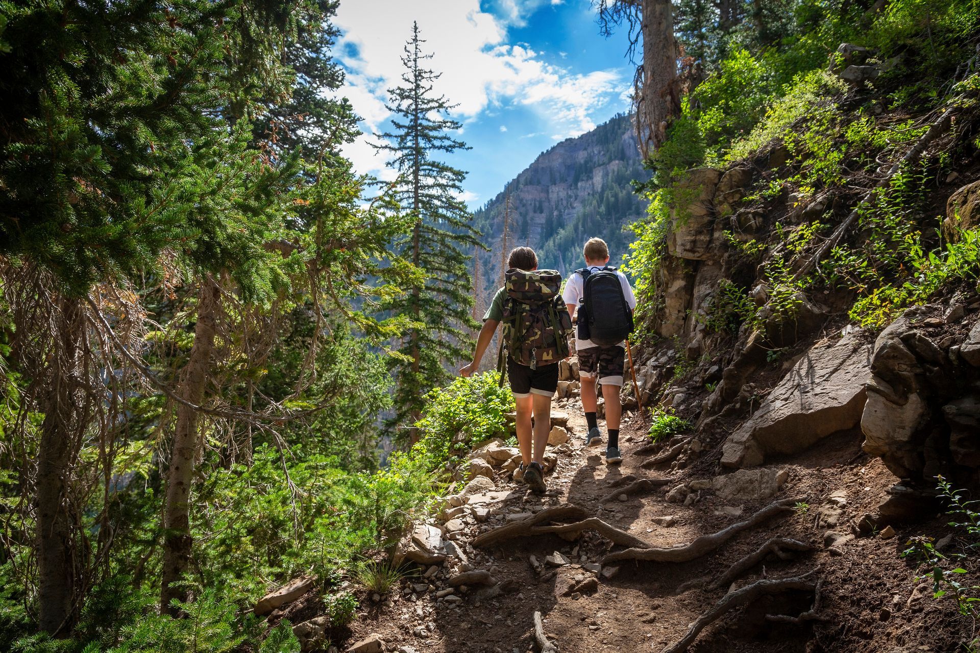 Two hikers on a narrow, dirt trail in a mountain setting. They are surrounded by green foliage, with a rocky cliff in the background.