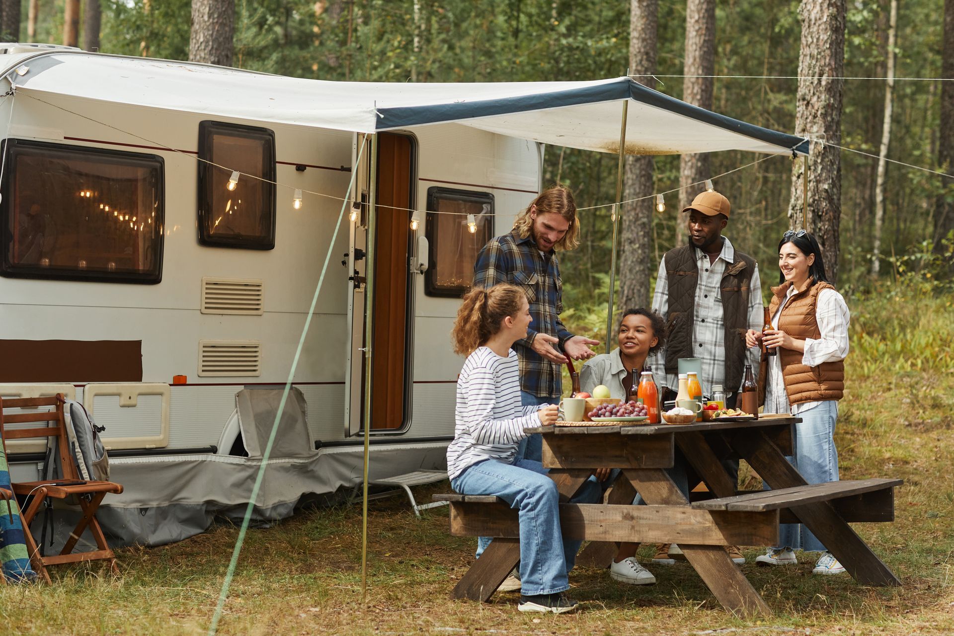 A diverse group enjoys a picnic at a campsite with a camper trailer. People eat, laugh, and socialize around a wooden picnic table in a forest setting.