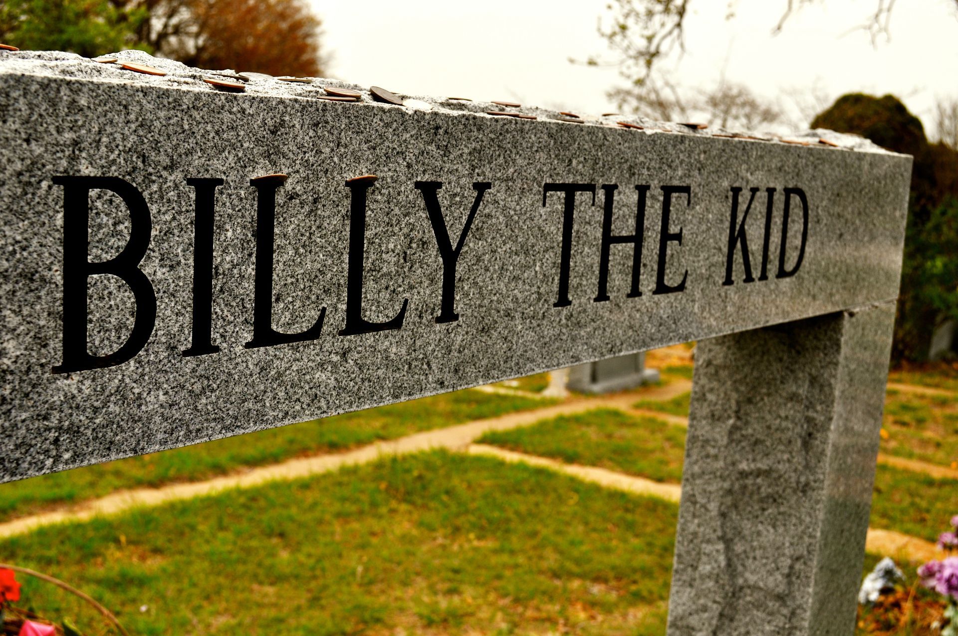 Billy the Kid's tombstone: gray granite marker with black text in a cemetery, surrounded by green grass.