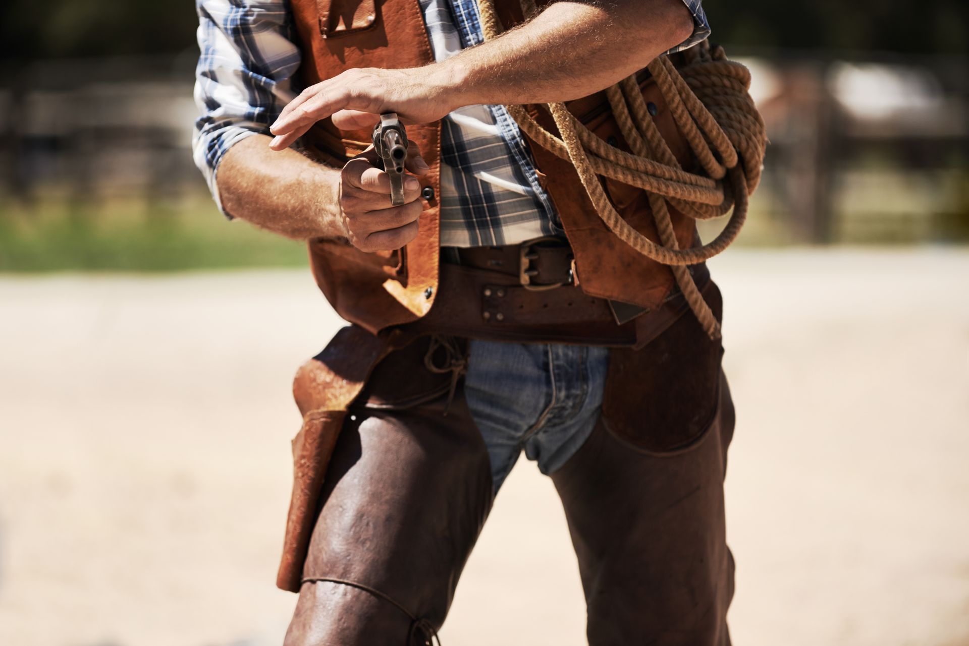 Cowboy adjusting gun, wearing leather vest, chaps, and carrying rope outdoors.