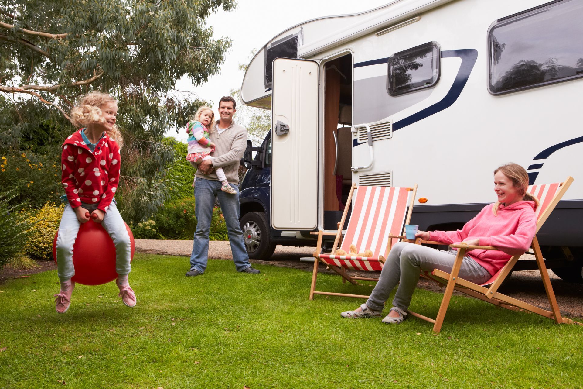 Family enjoying an RV vacation: girl bouncing on a red ball, parents relaxing outside, sunny day.