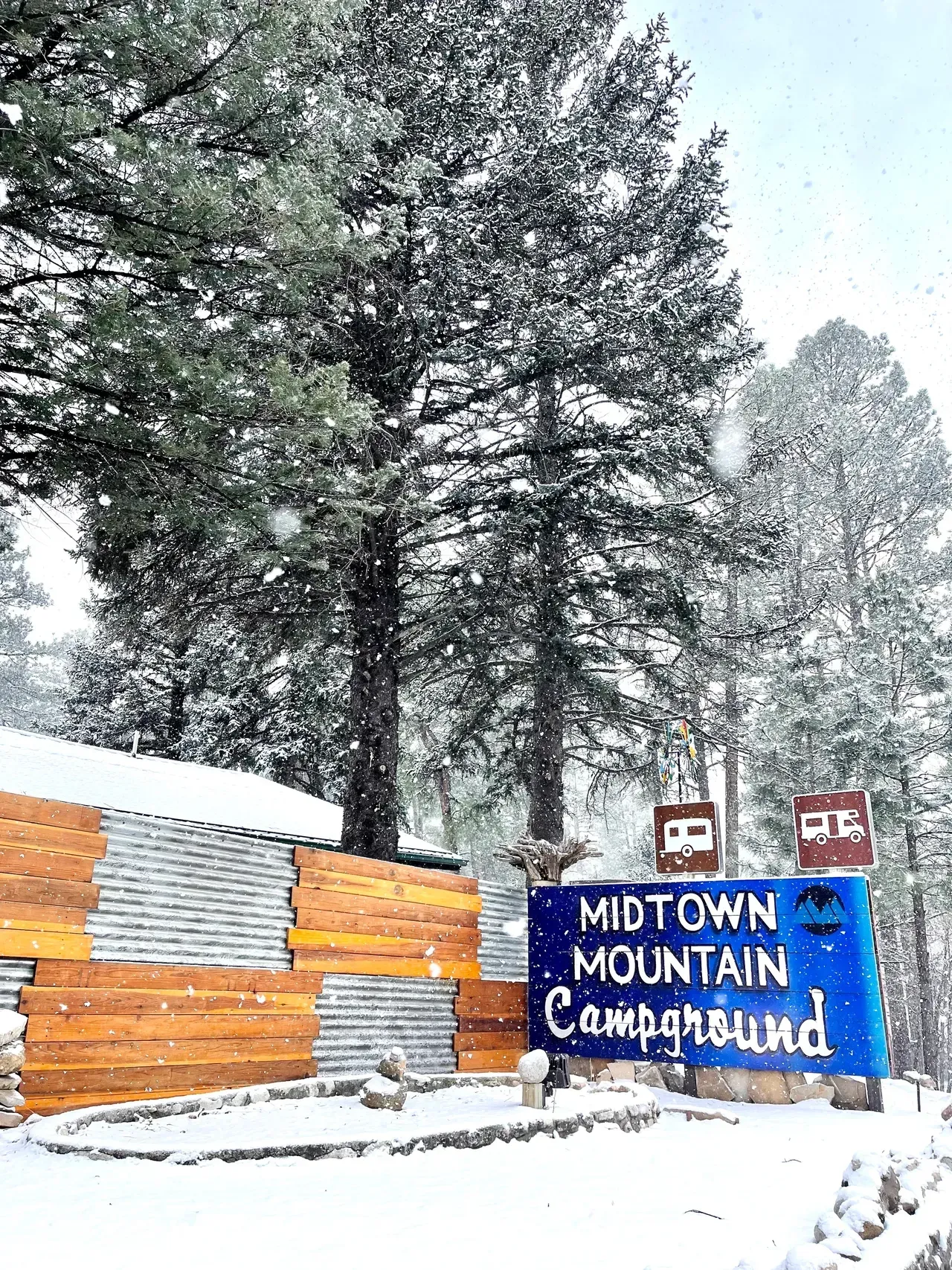 A blue sign for midtown mountain campground is surrounded by snow covered trees.