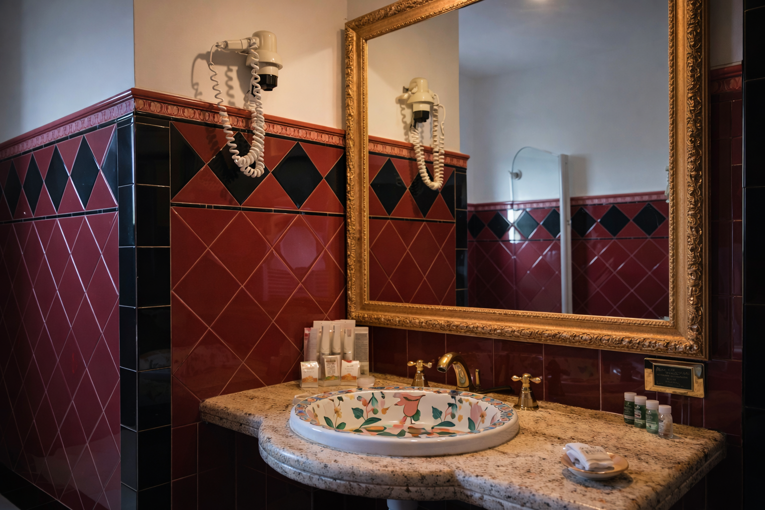 Bagno con specchio decorato, pareti piastrellate rosse e nere, lavandino decorato e piano di lavoro in granito.