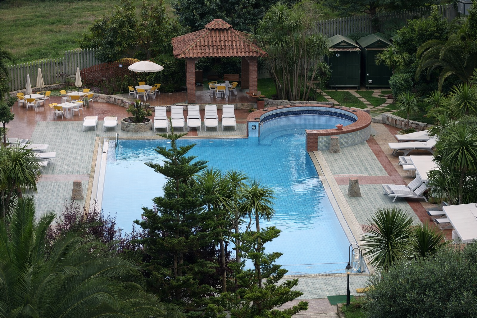 Piscina con sedie a sdraio, tavoli con ombrelloni e gazebo ombreggiato in un ambiente esterno.