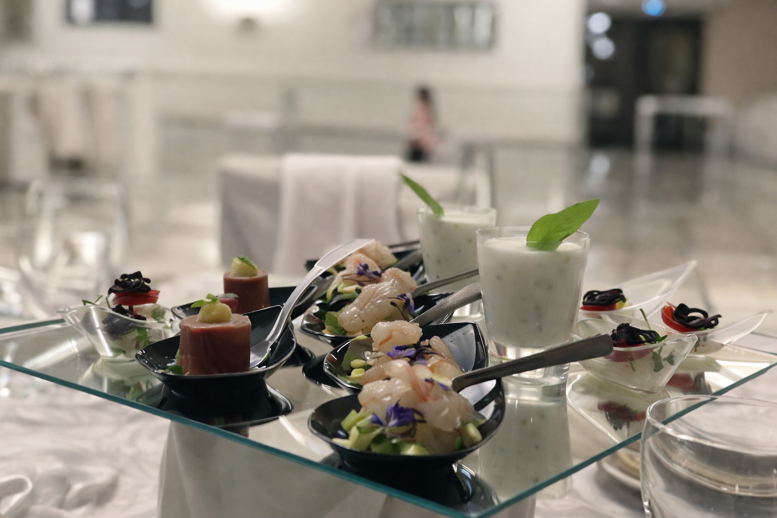 Antipasti su un vassoio a specchio in una stanza luminosa: piccoli piatti con frutti di mare, tonno e bevande cremose.