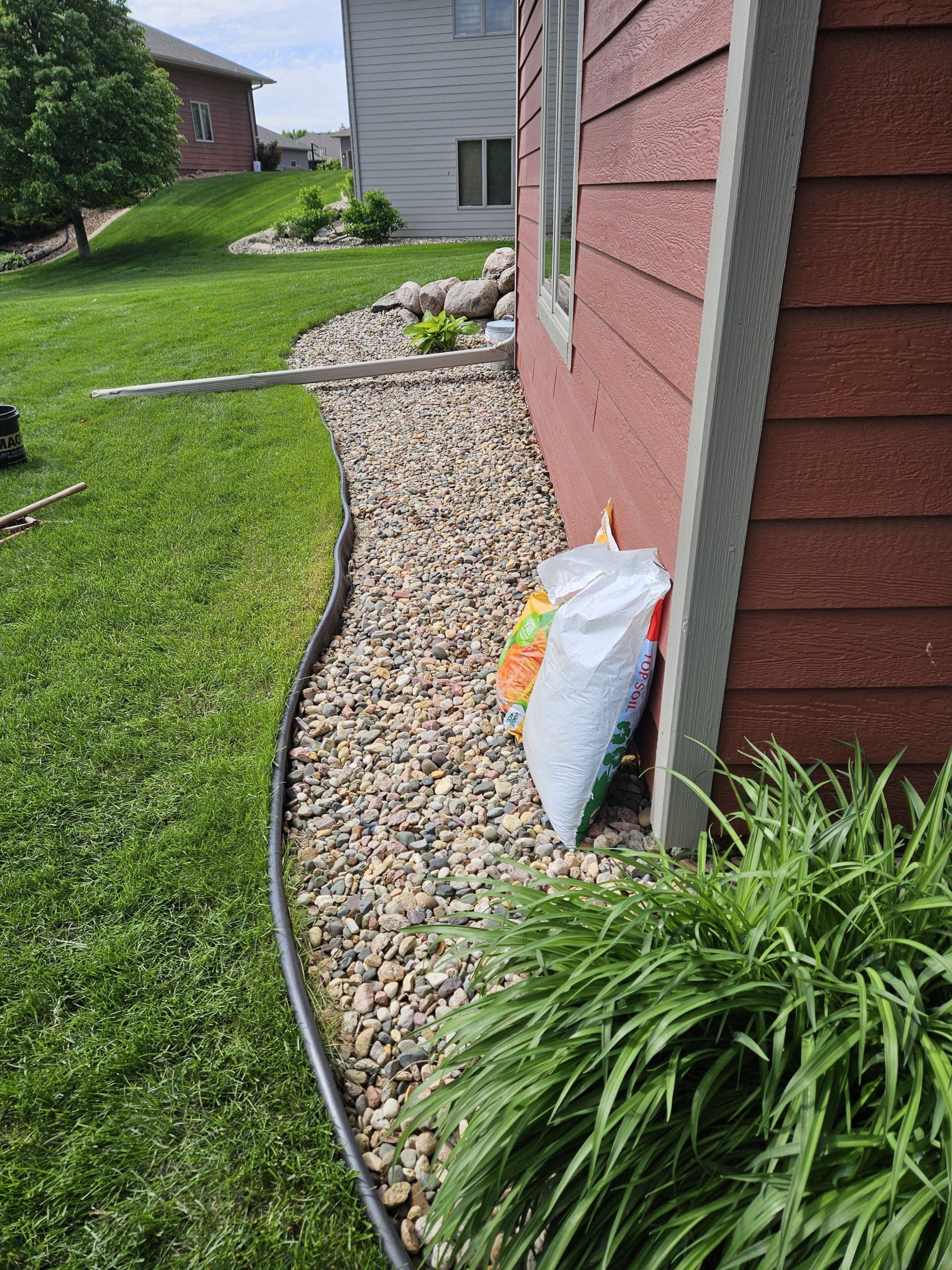 Side of a red house with a gravel bed and green lawn, bordered by a plastic edge. A bag rests against the wall.