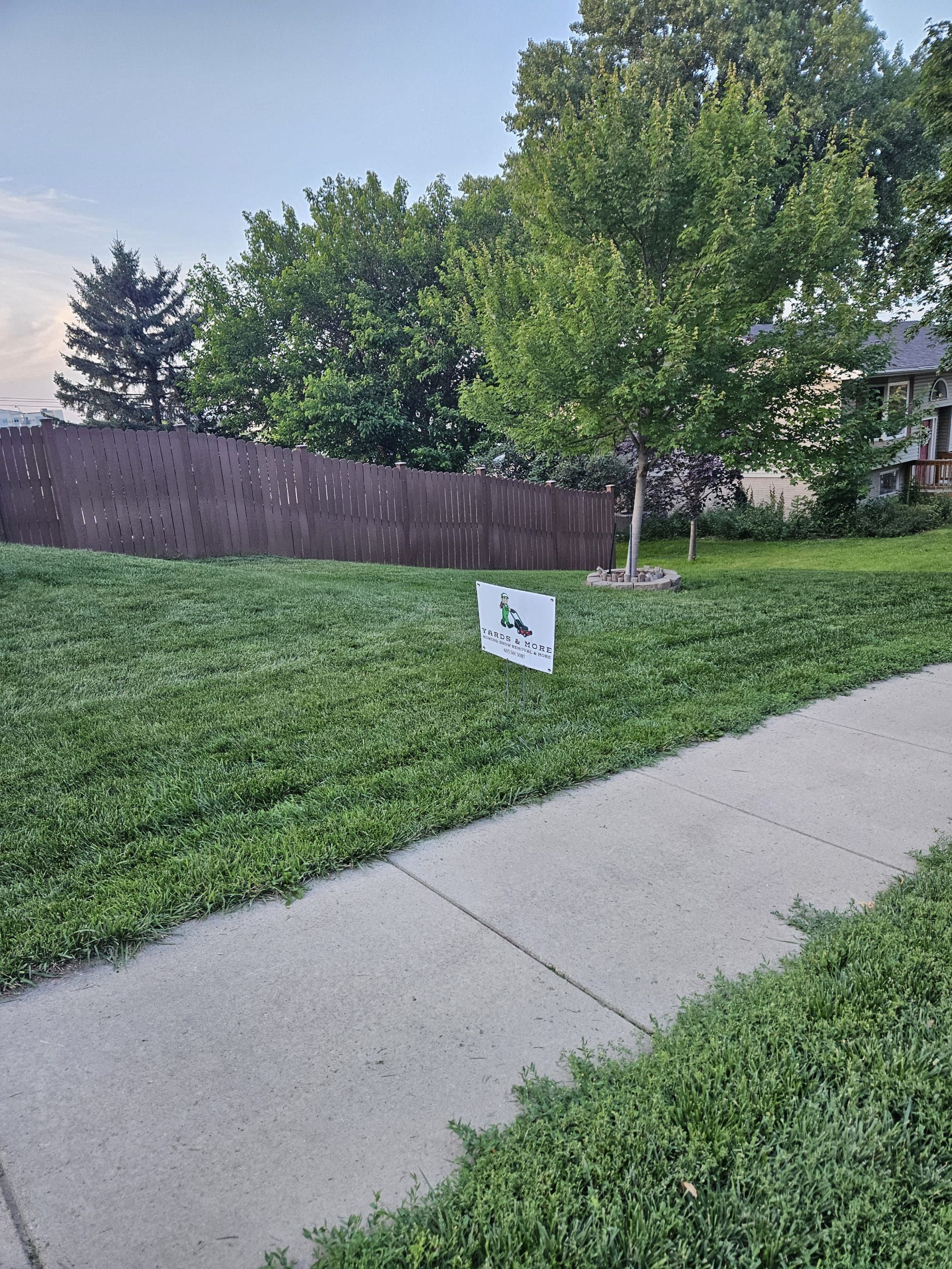 Grassy yard with sidewalk, brown fence, tree, and a sign.