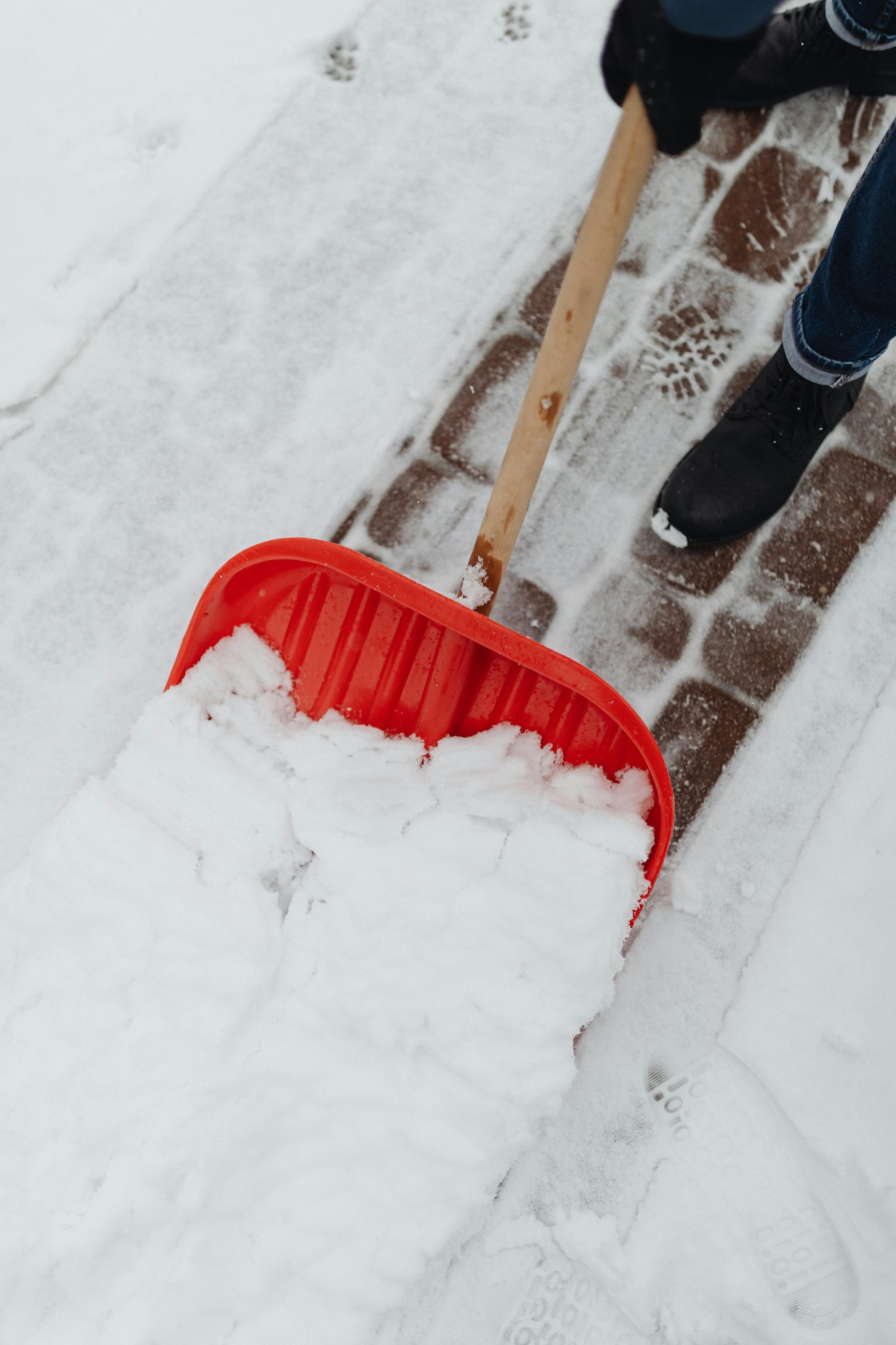 Person shoveling snow off a brick pathway with a red shovel, wearing gloves and boots.