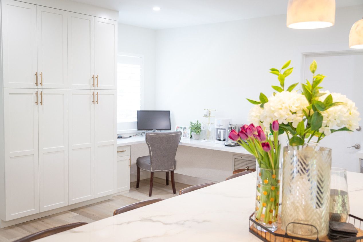 A kitchen with a desk and chair and a vase of flowers on the counter.