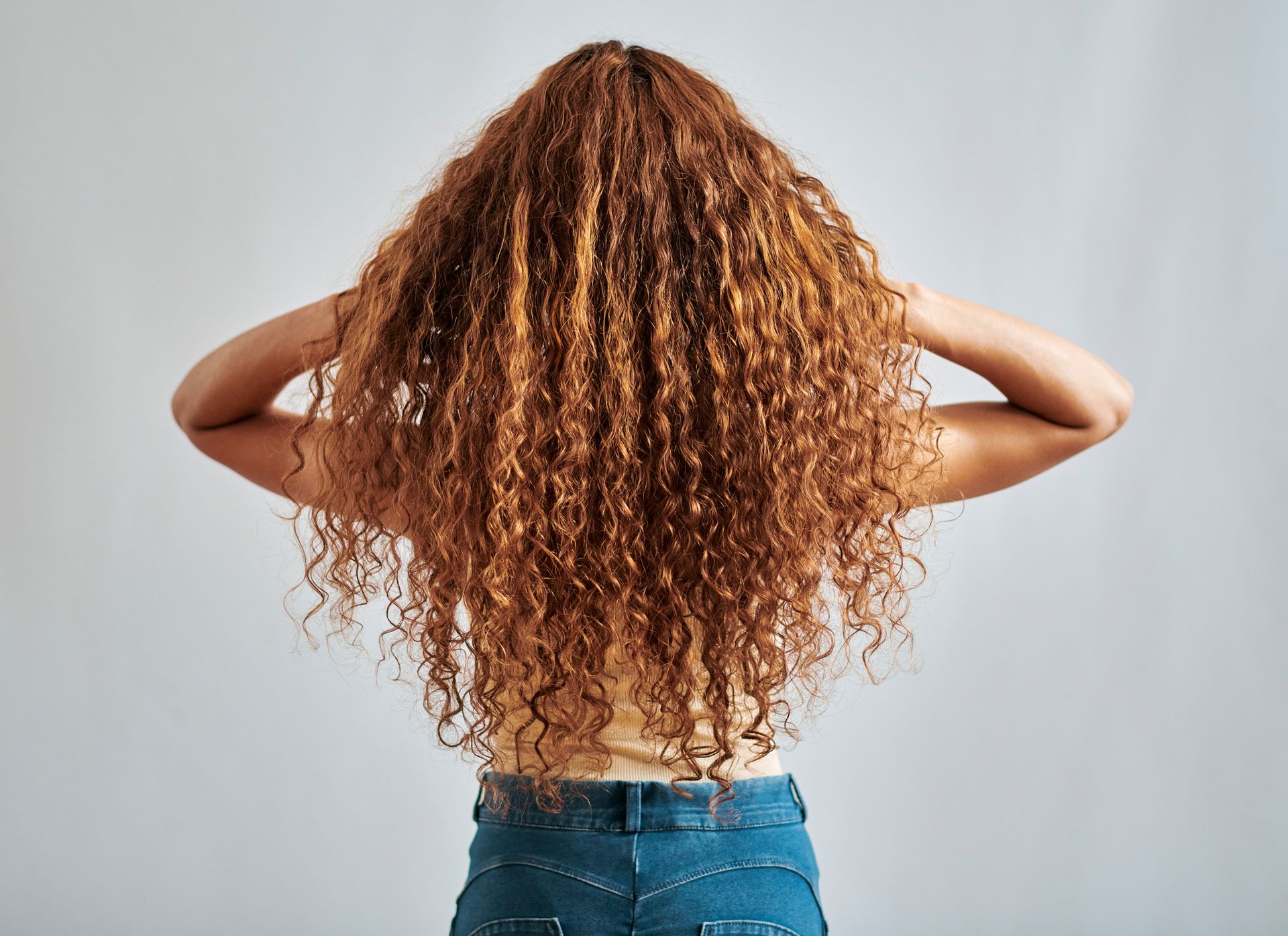 A woman with long curly hair is standing with her hands behind her head