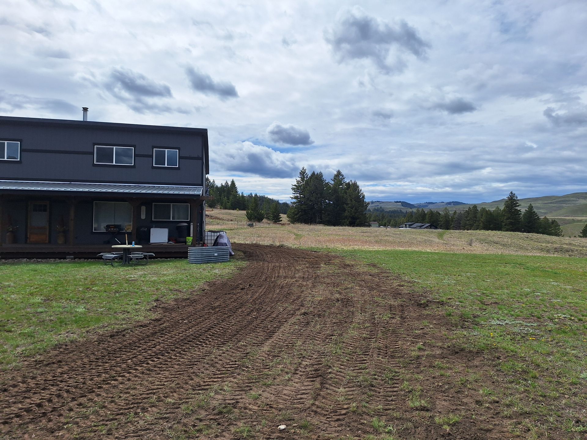 A two-story dark house on a grassy plot with a tilled area. Cloudy sky and distant hills are in the background.