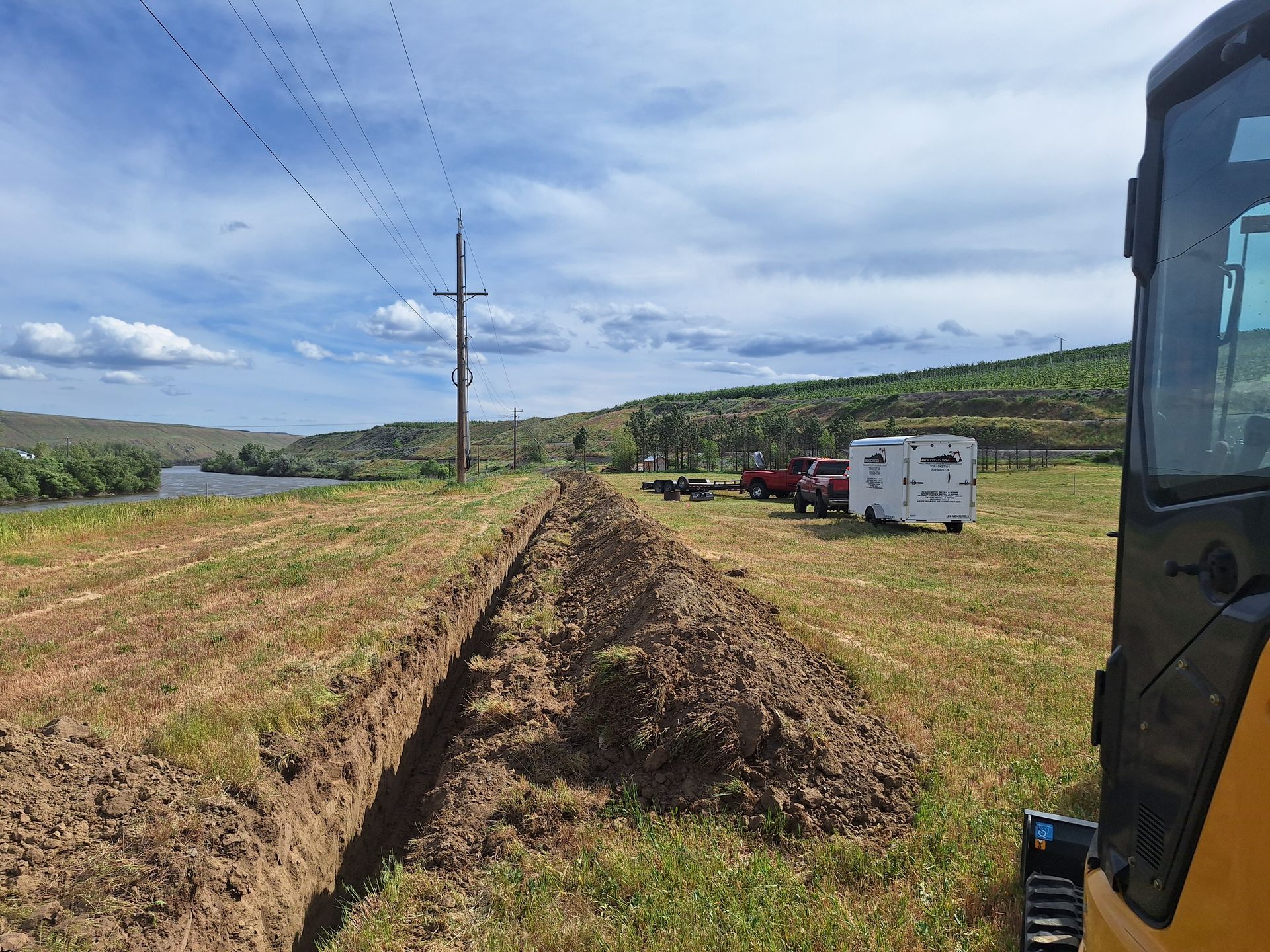 Trench dug in a grassy field near power lines, with trucks in the distance under a blue sky.