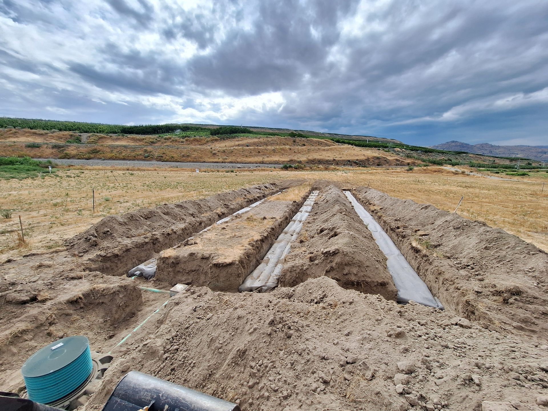 Trenches dug in dry dirt, possibly for a septic system installation. Cloudy sky, arid landscape.