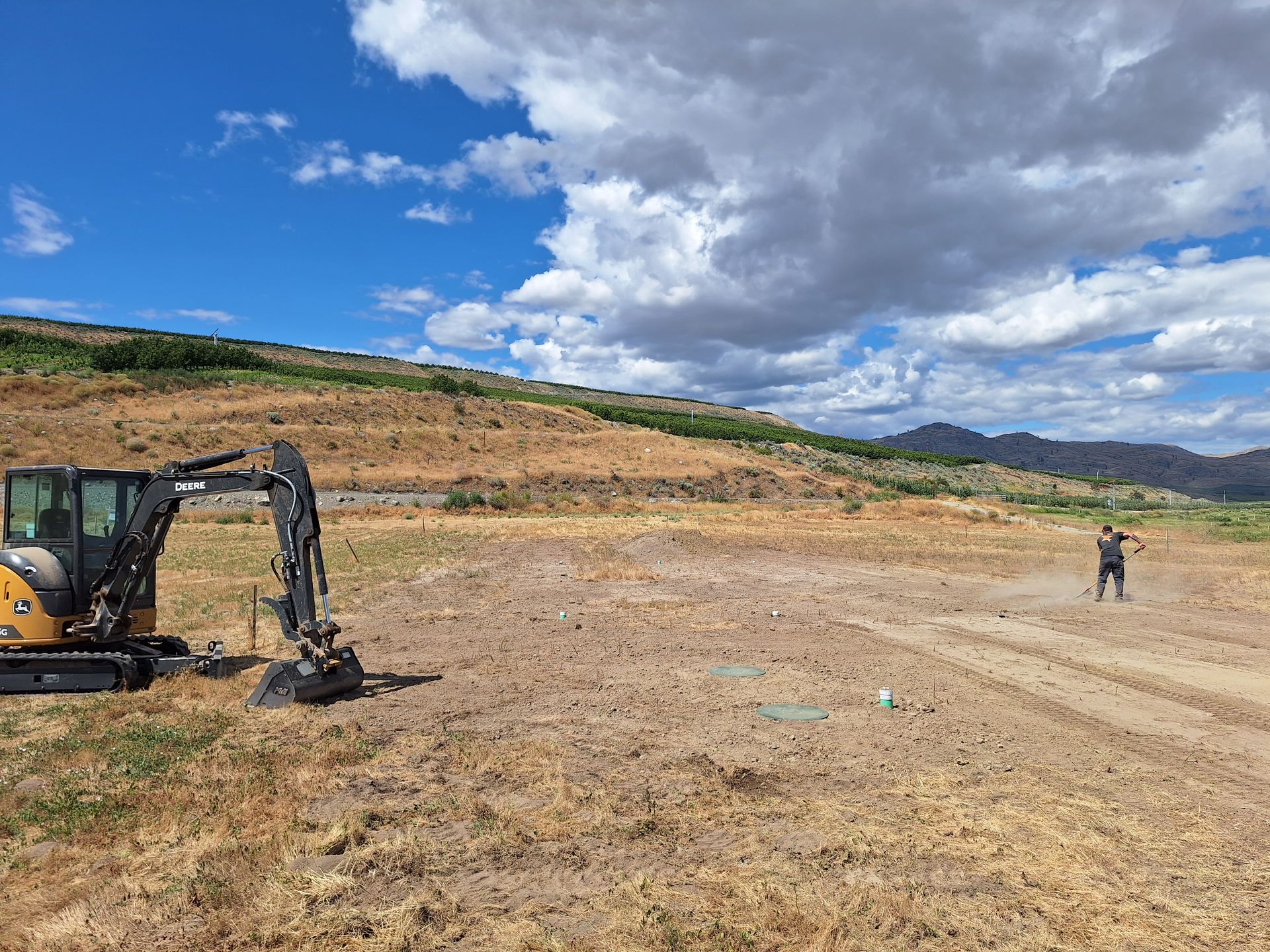 Construction site with small excavator, man surveying; dry field, hill, blue sky with clouds.