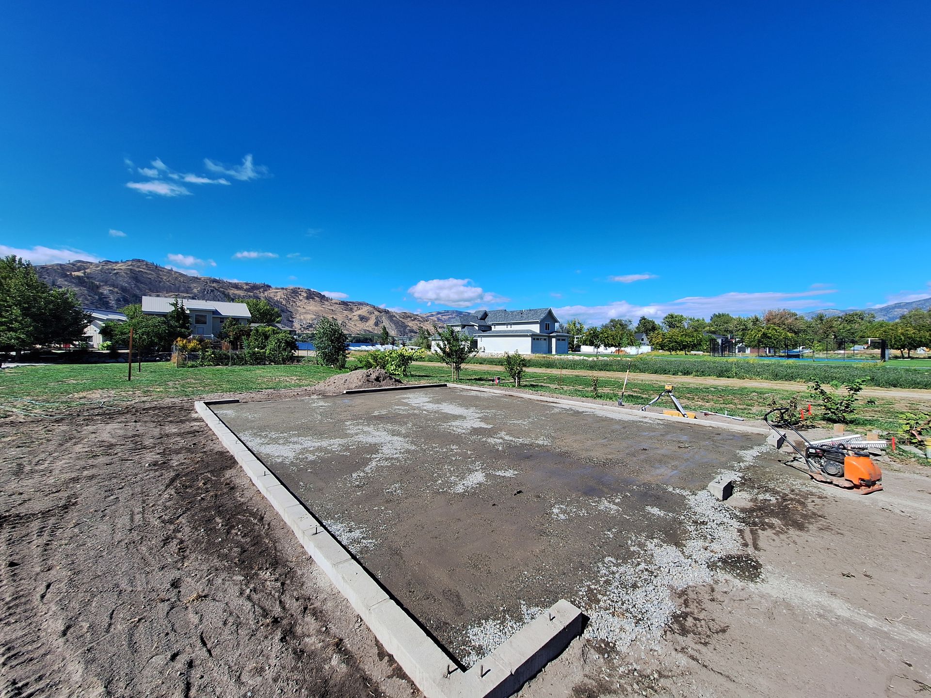 Construction site with foundation slab, dirt, clear blue sky, distant buildings, mountains.