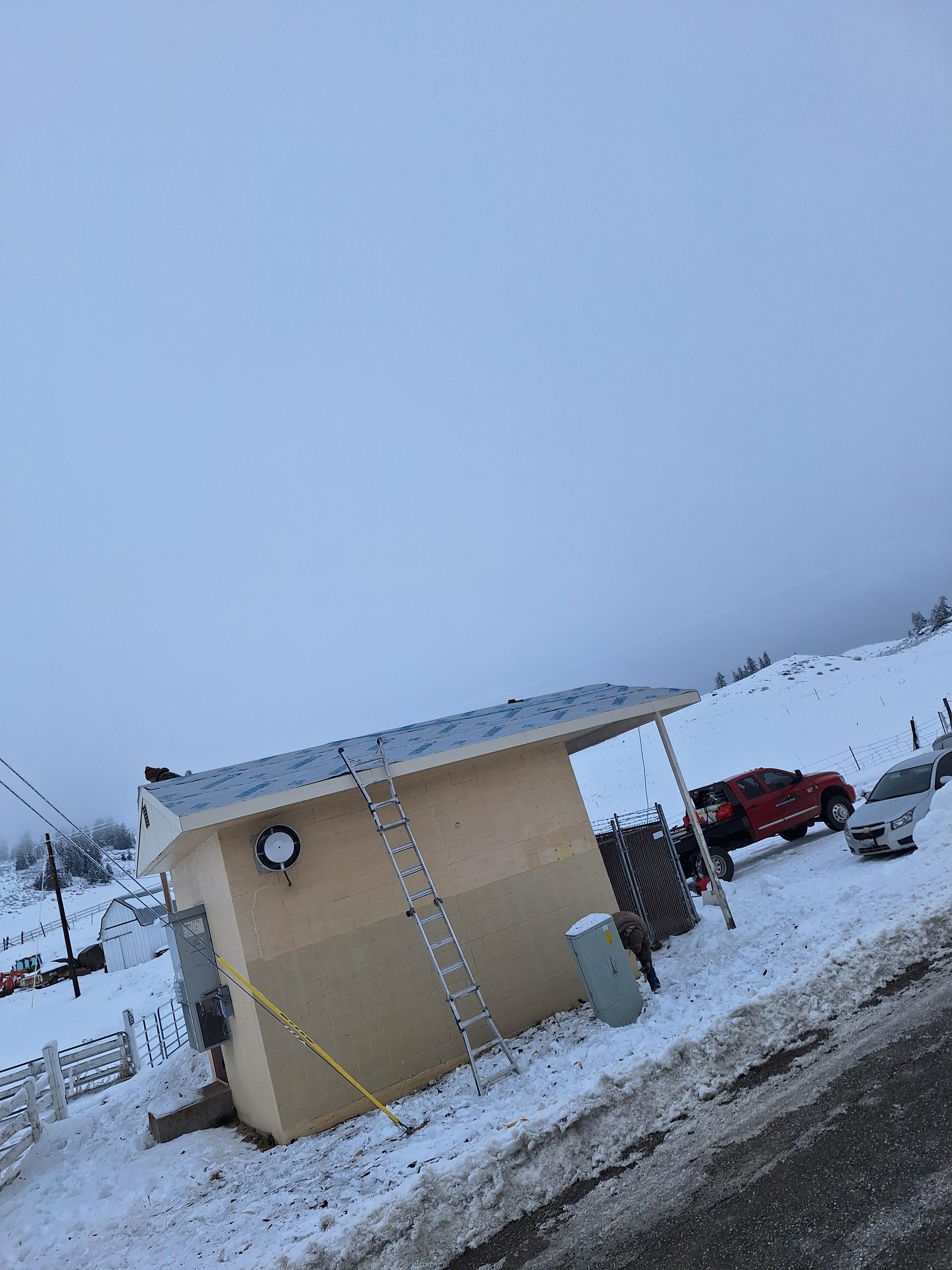 Small building on a snow-covered hillside. Red truck and car parked nearby. Overcast sky.