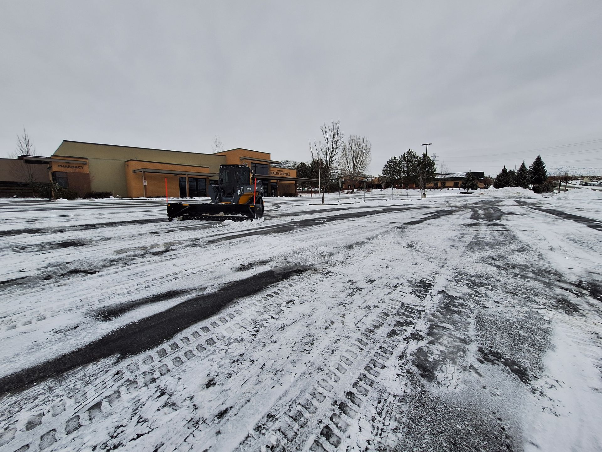 Snow-covered parking lot with a yellow snowplow clearing a path in front of a tan building under an overcast sky.