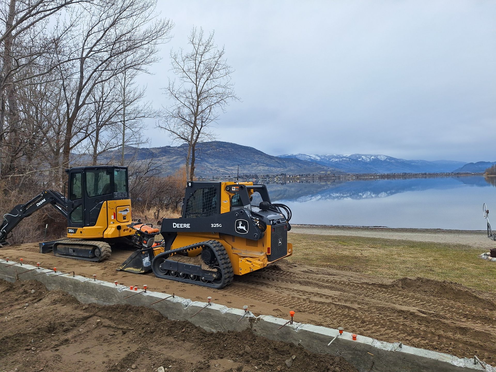 Construction site with a yellow skid steer and small excavator near a lake. Cloudy sky, mountains in the background.