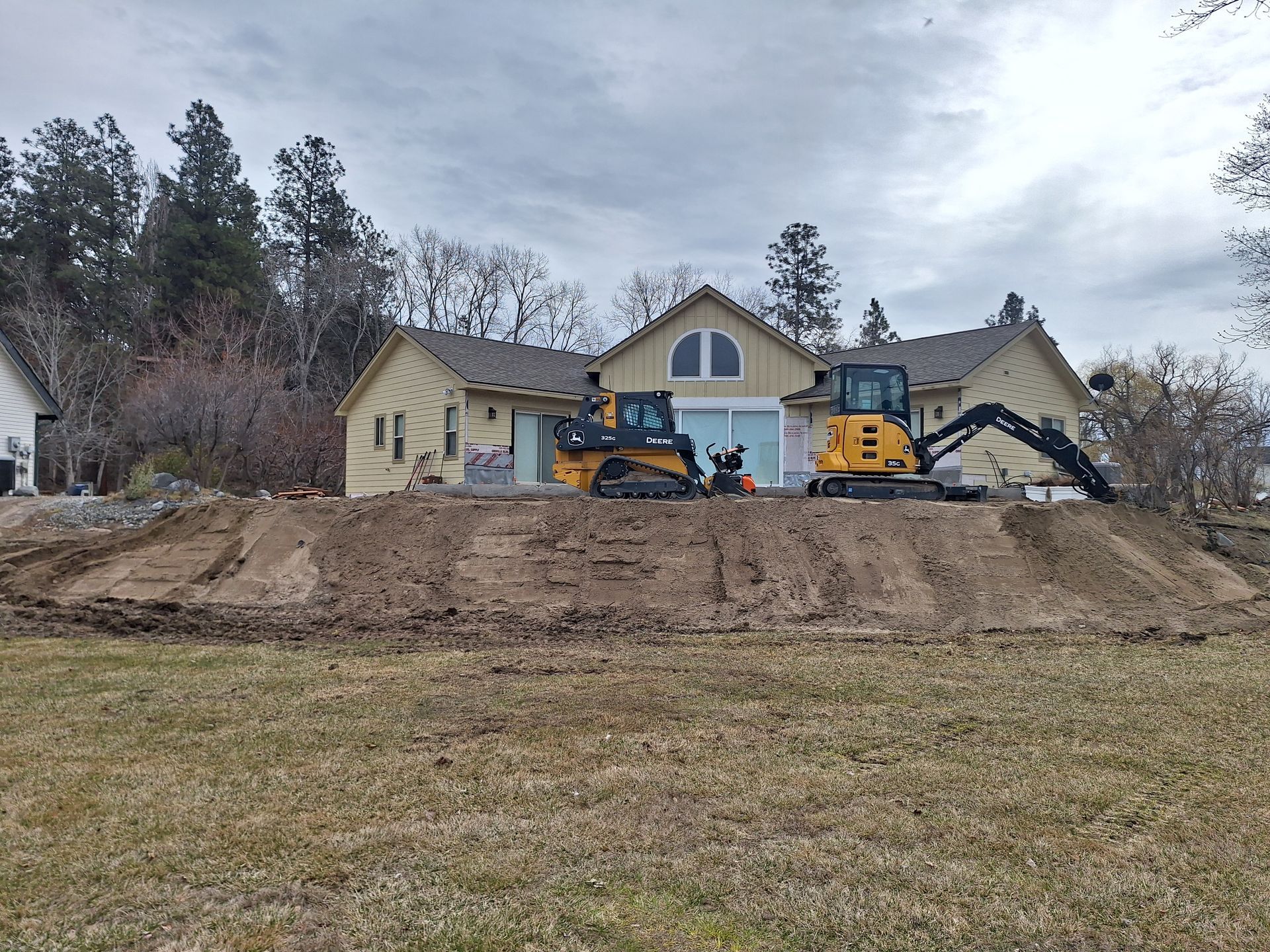 Two excavators in front of a light yellow house with construction debris and a cloudy sky.