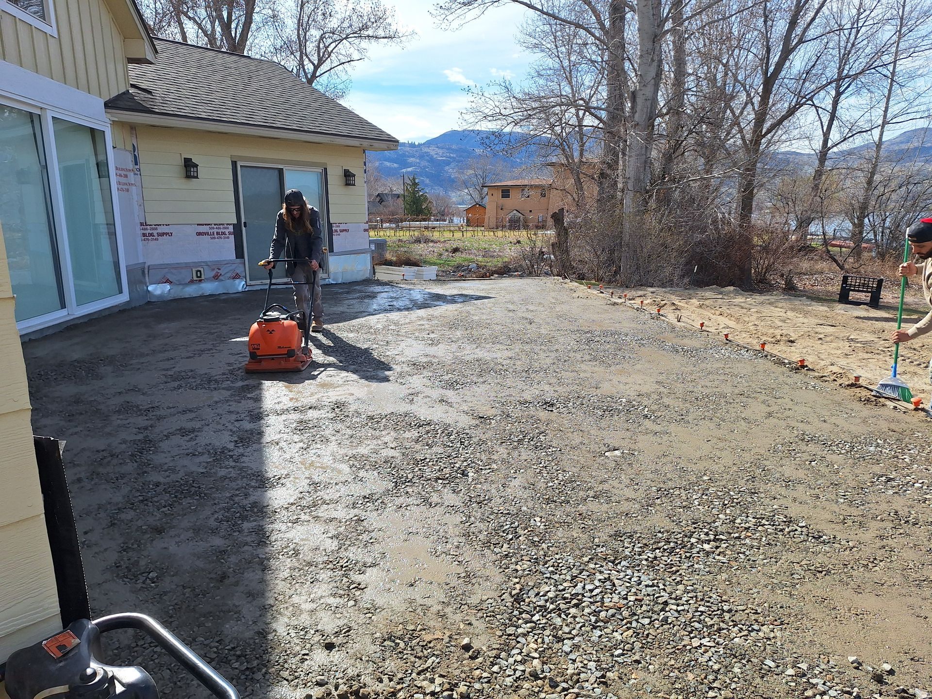 Workers compacting gravel patio outside a yellow house. One uses a compactor, another sweeps.