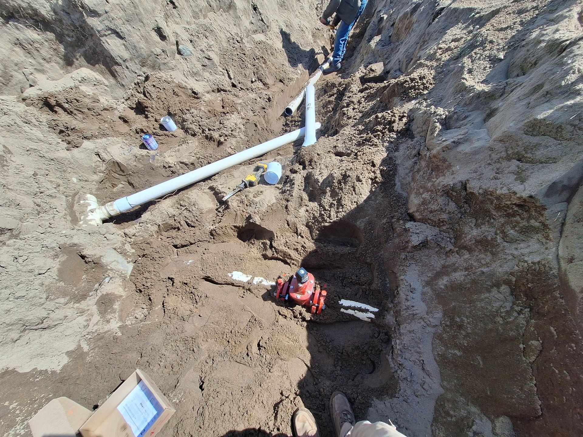 Trench with blue and white pipes, fittings, and a red valve, in dirt.
