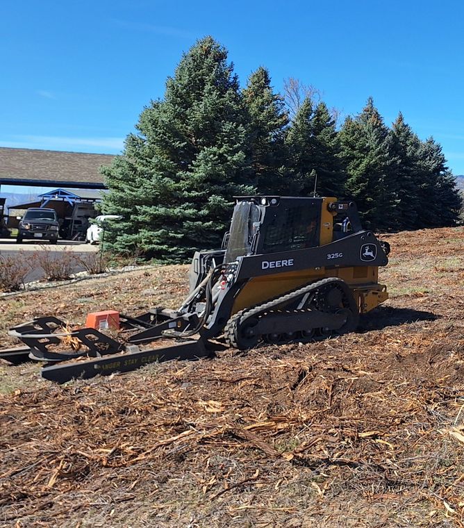 A small, tracked skid steer with a mulcher attachment on a brown mulch pile, in front of evergreen trees.
